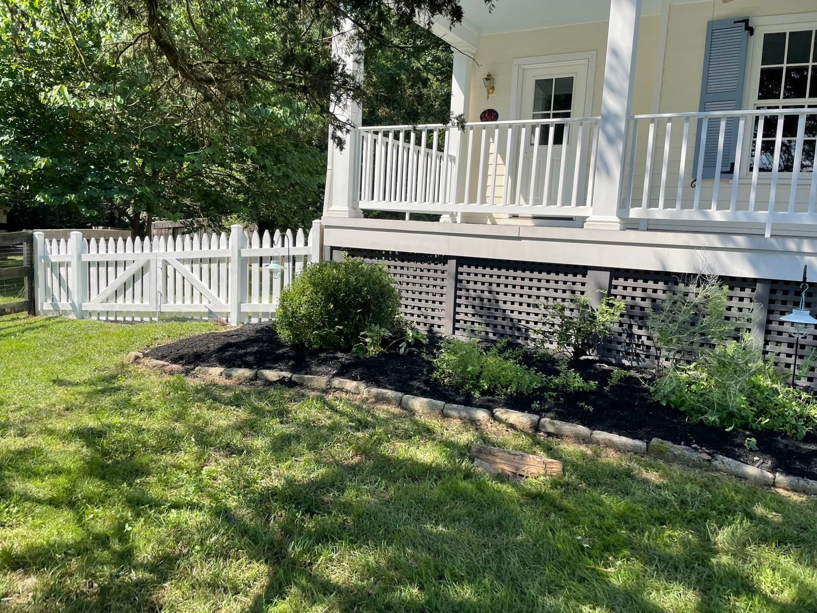 White house porch with railing and lattice, bordered by shrubs and a grassy yard.