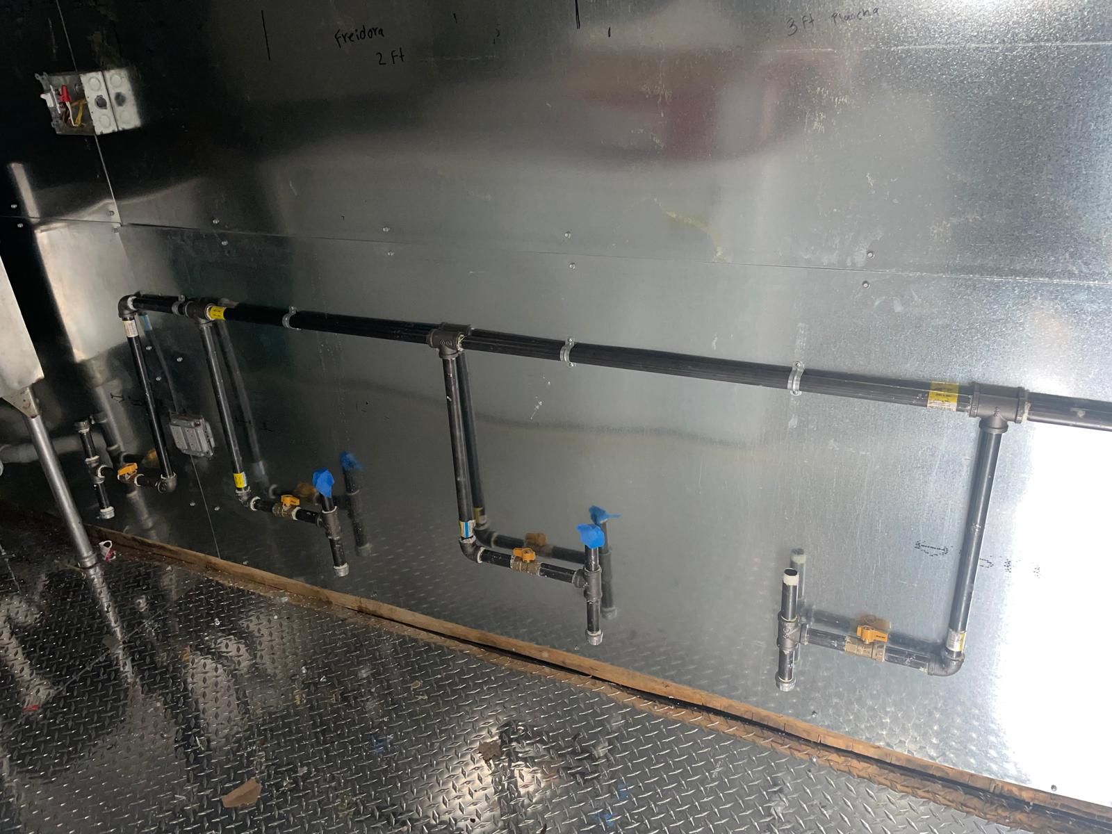 Black plumbing pipes and valves mounted on a metal wall in an industrial room.