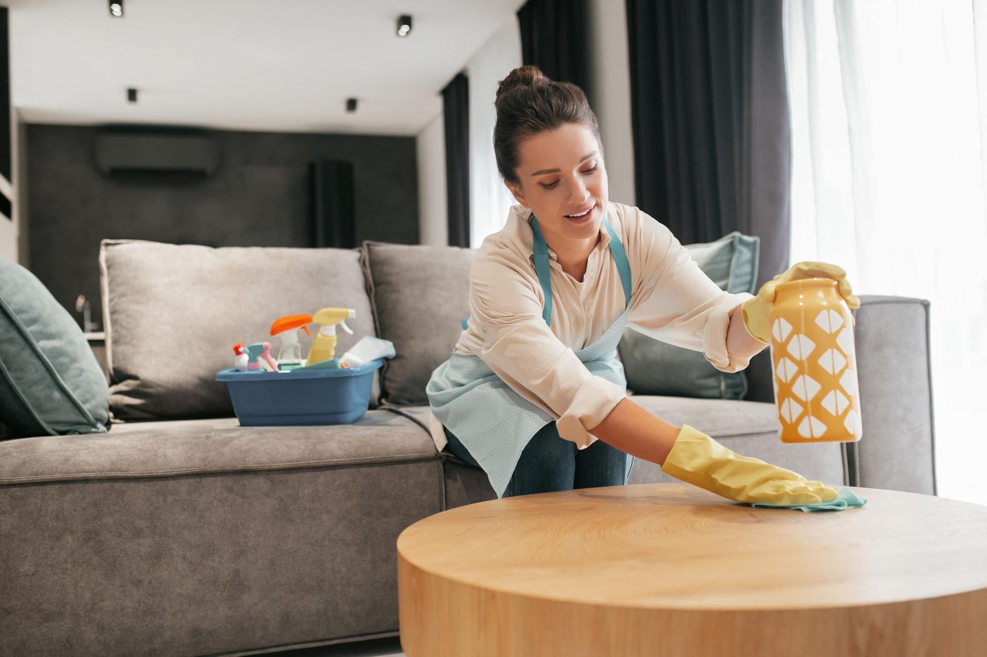 Woman cleaning a wooden coffee table in a living room, wearing gloves and an apron, with a vase.