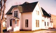 White house with dark roof and bay windows on a street corner, lit by warm sunlight