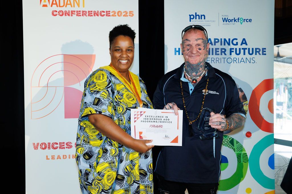 Two people smiling while standing in front of branded conference banners, holding a framed certificate at an event.