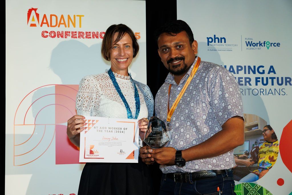 A woman and a man holding an award and a certificate in front of a conference banner with blue and orange branding.