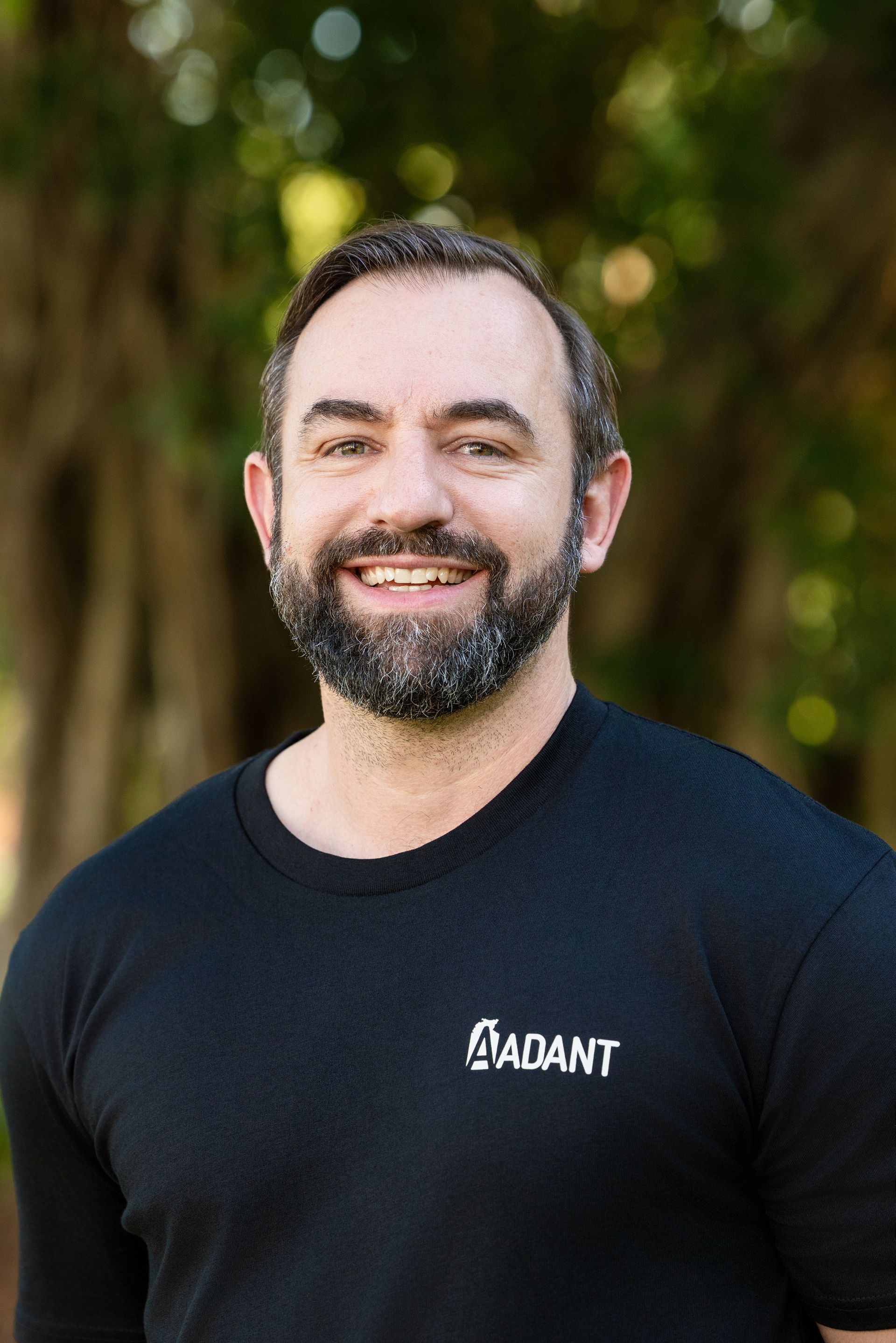 A smiling person with a beard wearing a dark sweater, positioned in front of a leafy green tree.