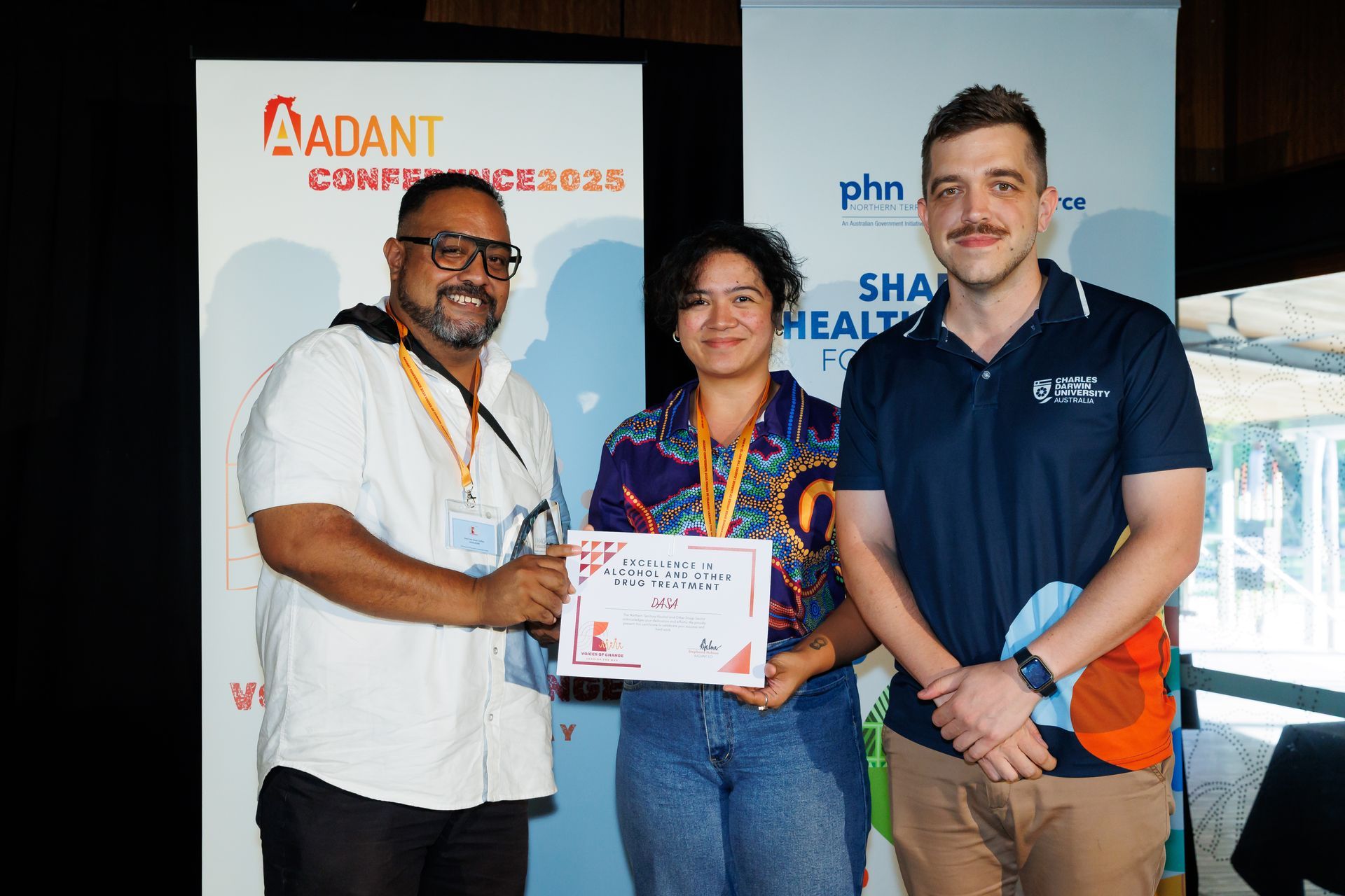 Three people stand smiling in front of banners for the ADANT conference, holding a certificate between them.