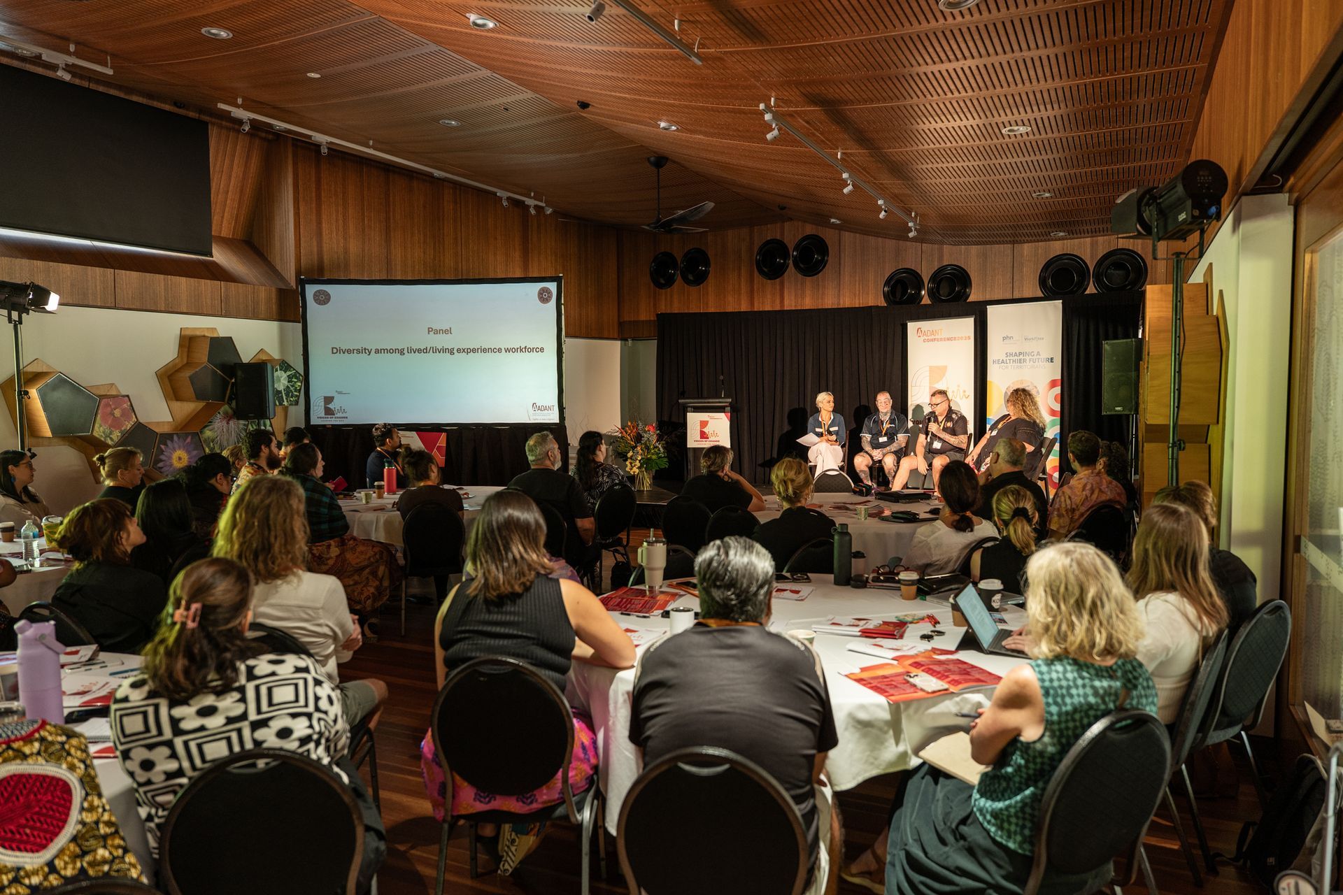 Attendees sit at round tables in a wood-paneled room, watching a panel discussion on stage under a projected screen.