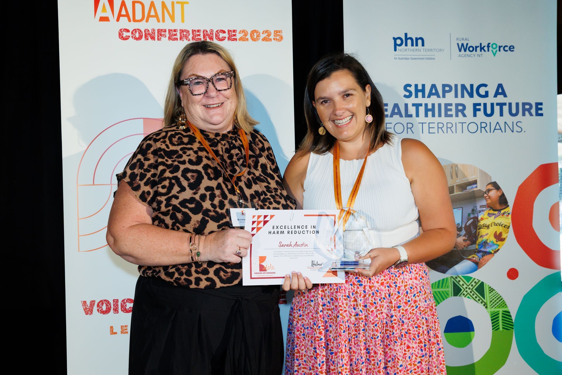 Two individuals standing in front of conference banners, holding a certificate and award during an event.
