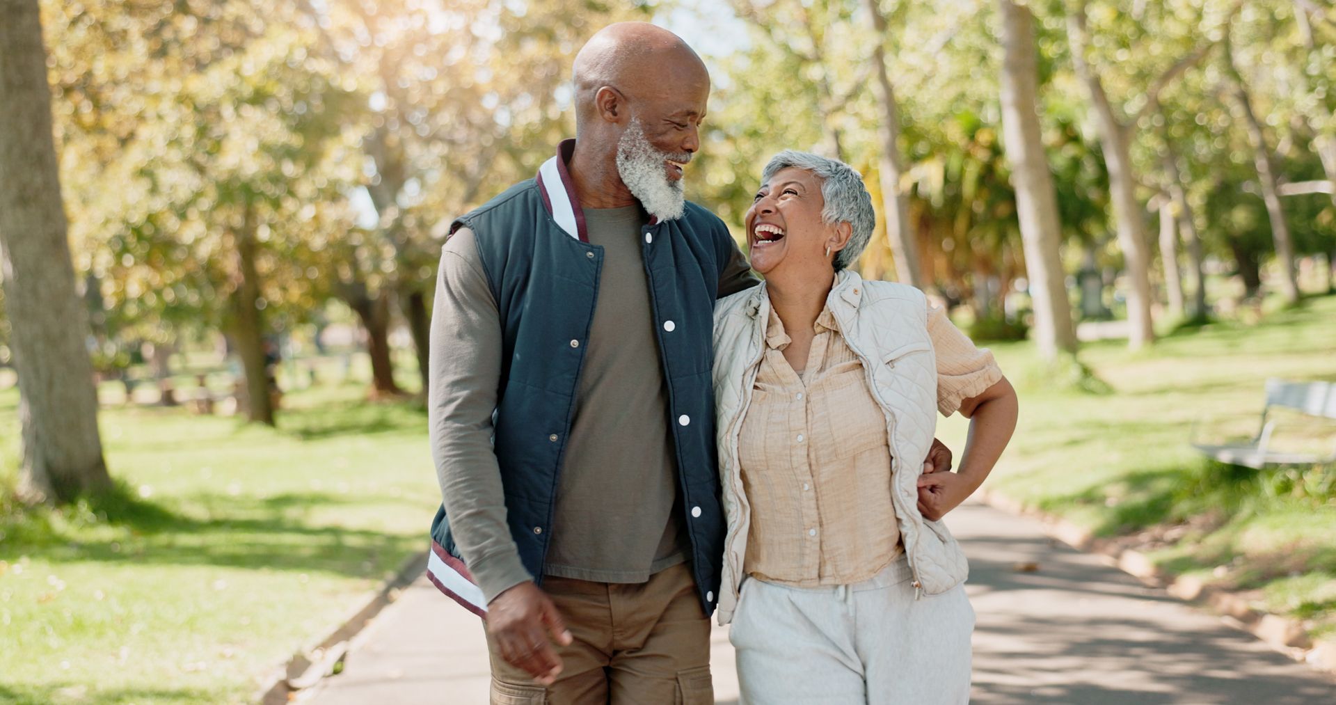 Two people walking arm-in-arm on a sunlit park path, smiling and laughing