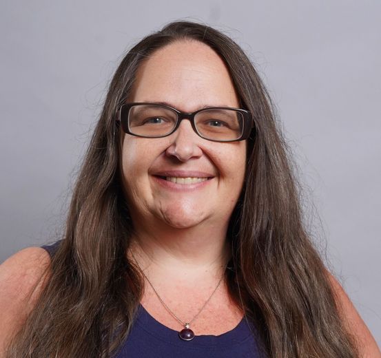 Smiling person with long dark hair and glasses, wearing a purple top against a gray background