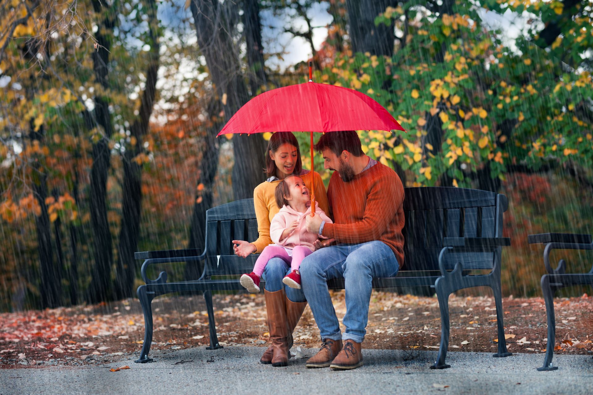 Couple and child sitting on a bench under a red umbrella in the rain at a park