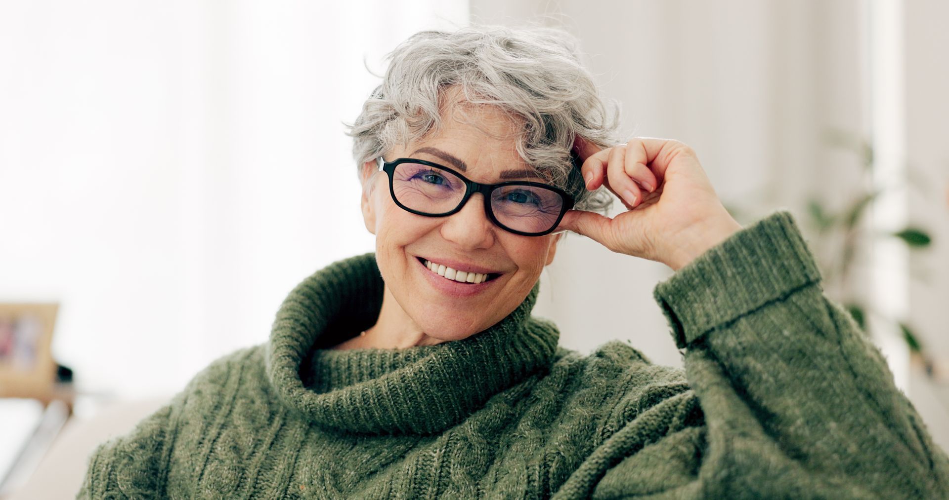 Smiling person in a green sweater adjusting glasses indoors near a bright window