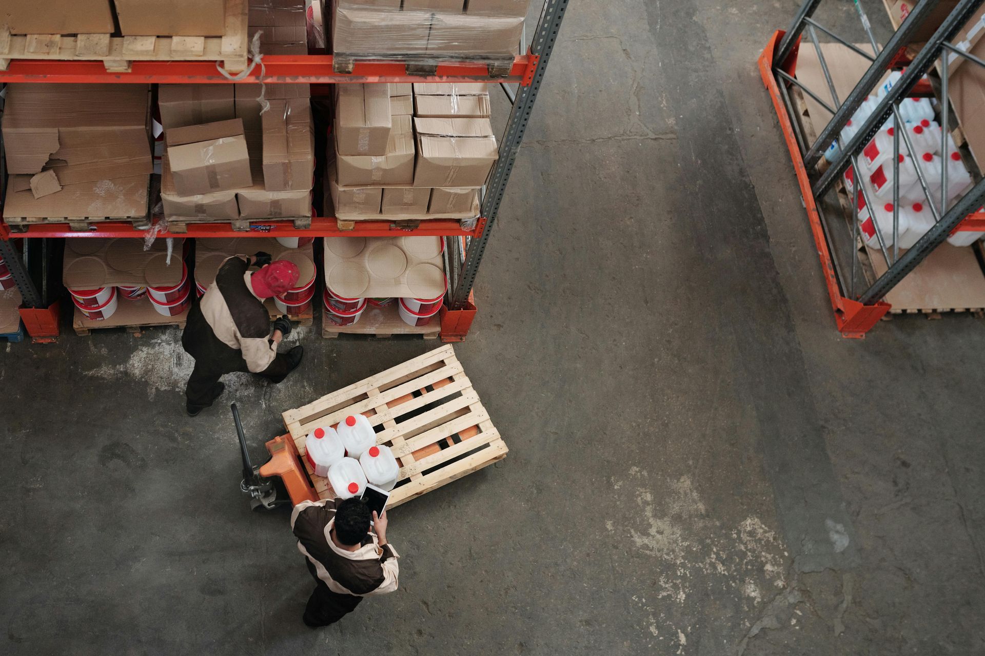 Two workers in a warehouse carrying a wooden crate past tall shelves of boxes and supplies.