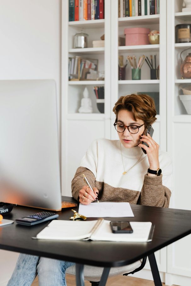 Woman writing at a desk while talking on the phone in a bright home office with bookshelves.