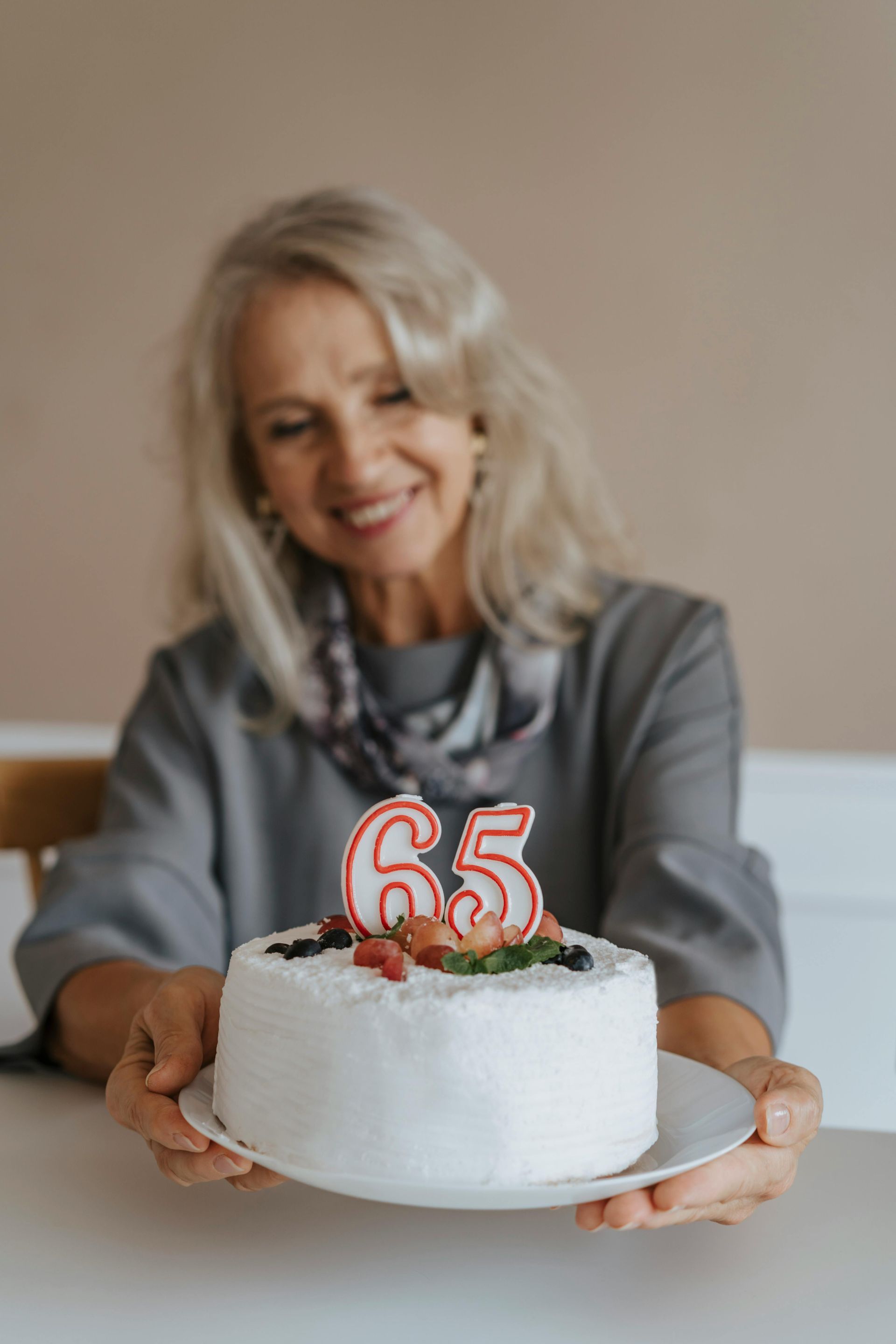Woman holding a white cake with a red “65” candle, smiling indoors.