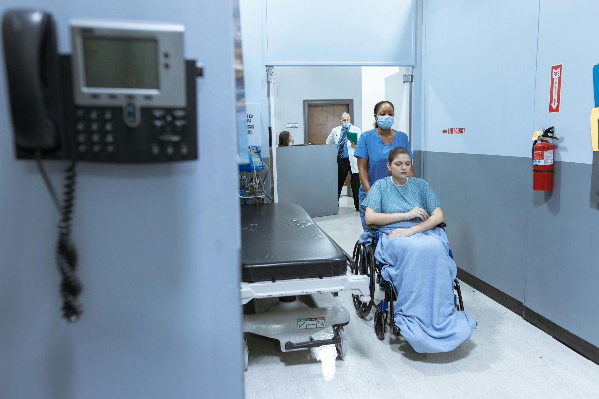 Patient in wheelchair being pushed down a hospital hallway by a masked caregiver.