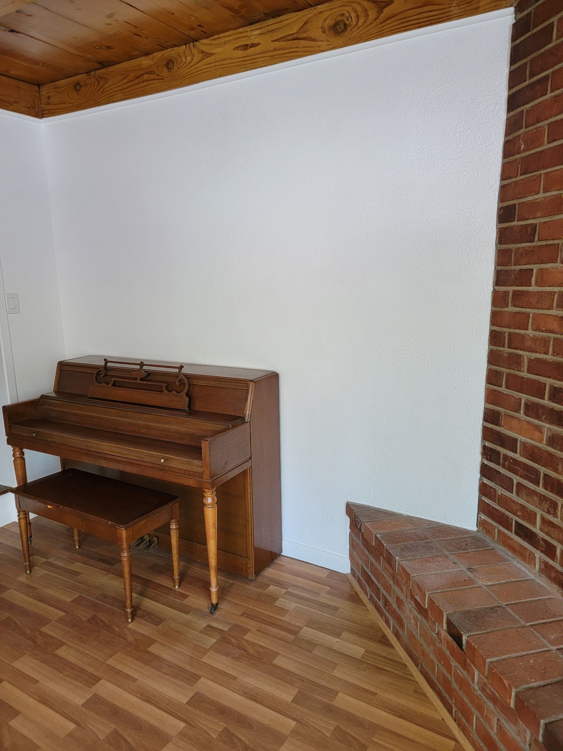 Small room with a wooden piano and bench against a white wall, brick accents, and wood floor.