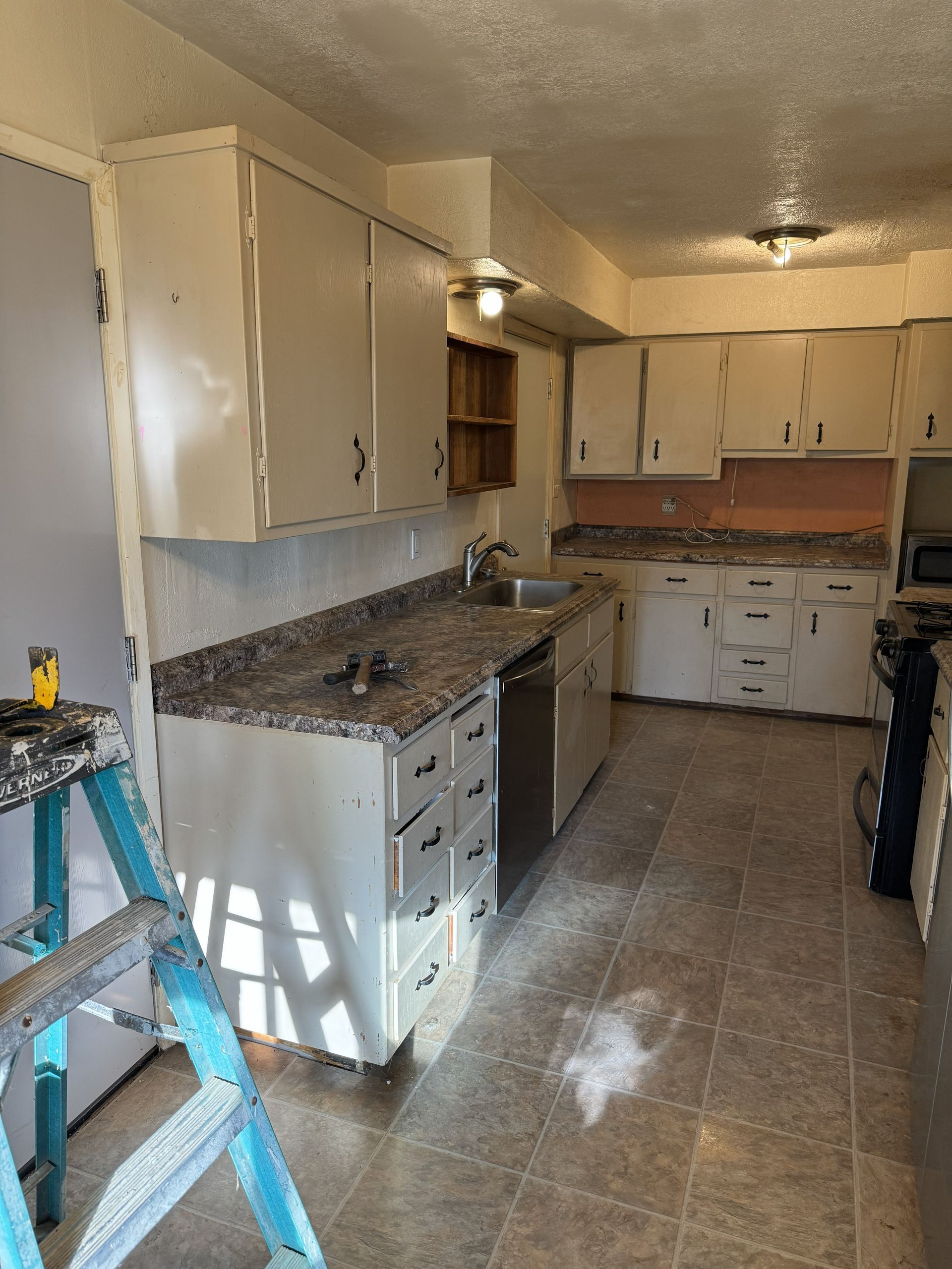 Small kitchen with white cabinets, granite counters, tiled floor, and a blue step ladder in the foreground
