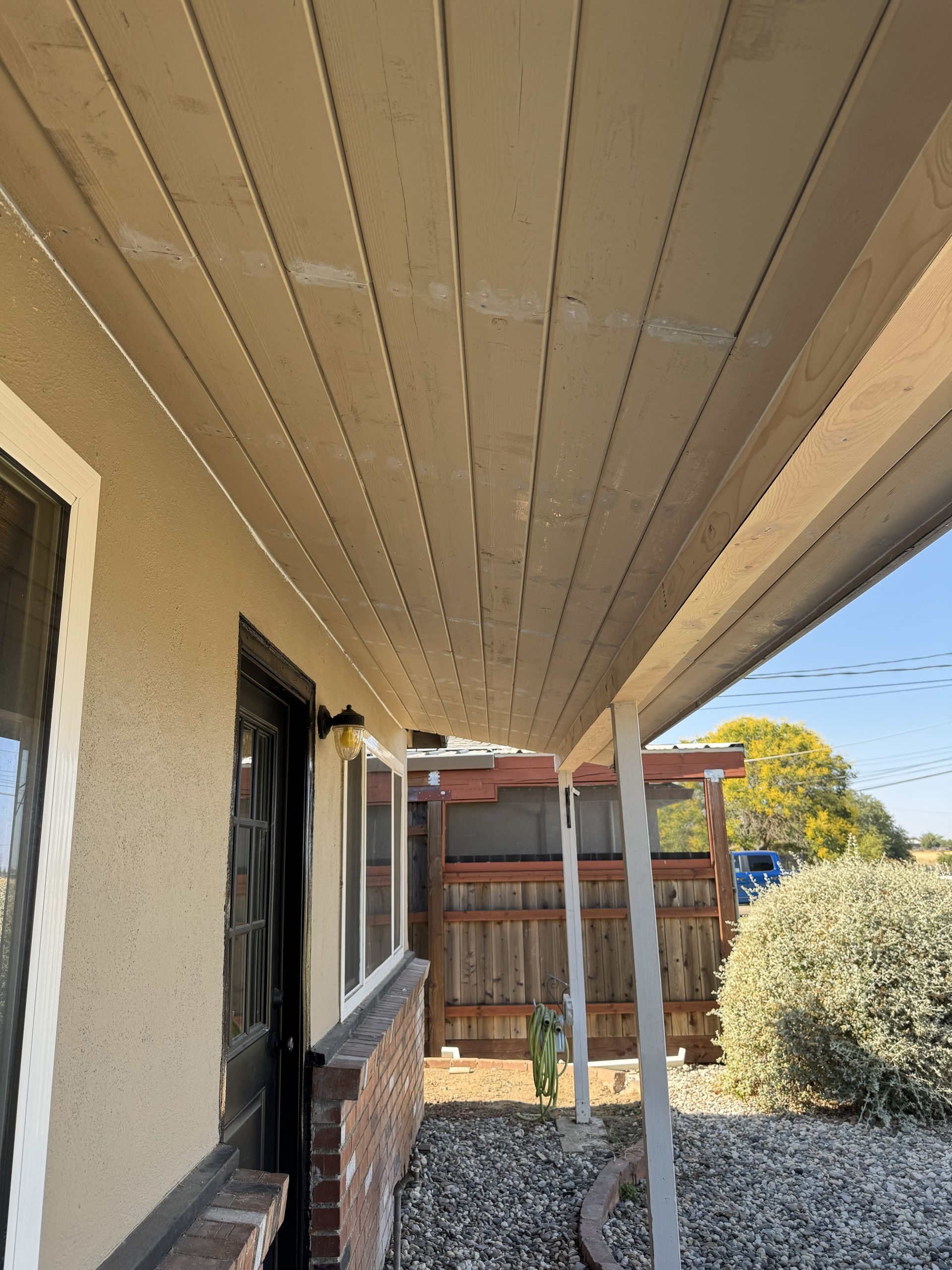 Covered backyard walkway beside a house, with beige siding, fence, and shrubs in sunlight