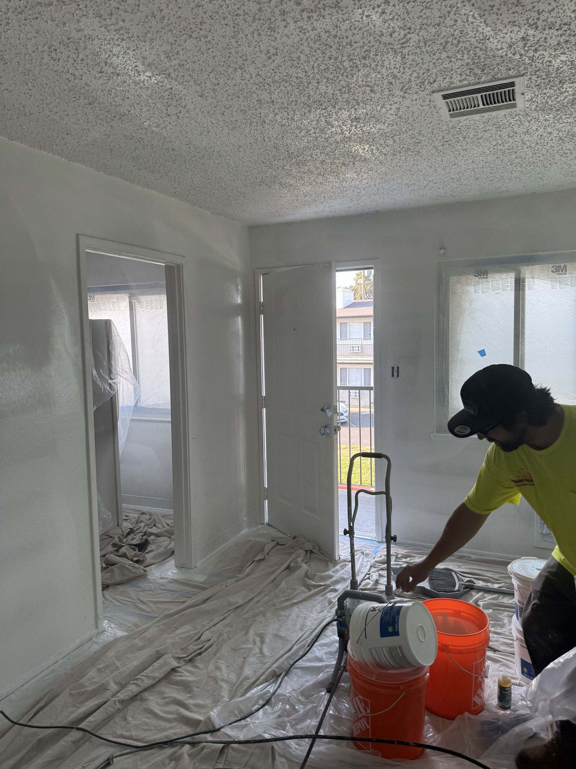 Renovation room with plastic-covered floor, white walls, and a worker sanding near buckets and a vacuum