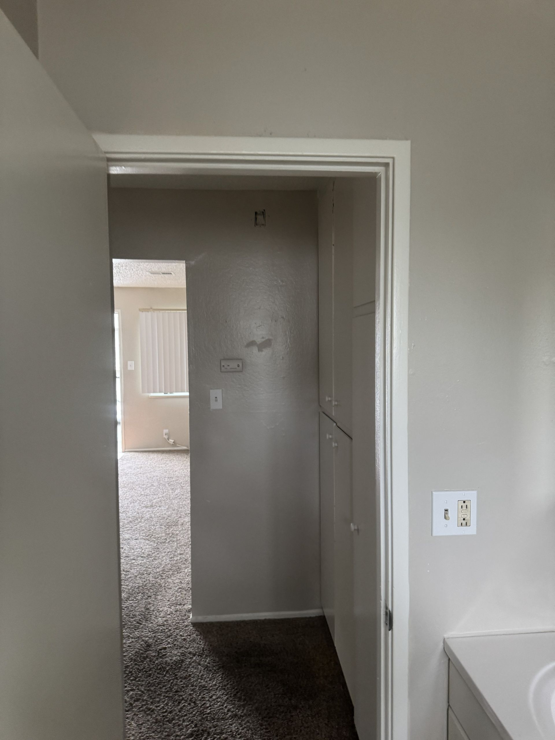 Empty hallway with white walls, doorways, and carpet leading to a room with vertical blinds.