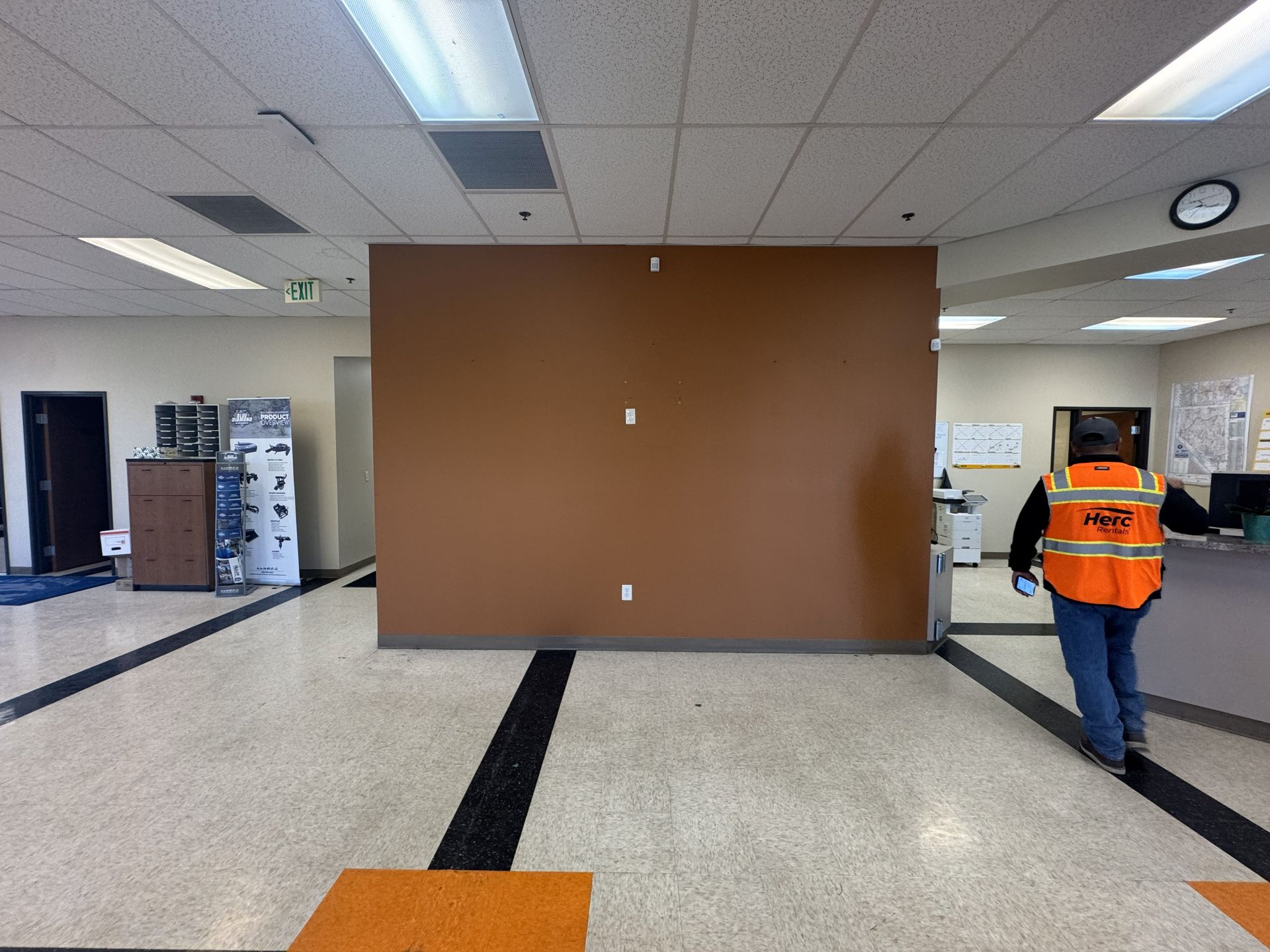 Large brown panel in a bright indoor lobby, with a worker in an orange vest walking on the right.