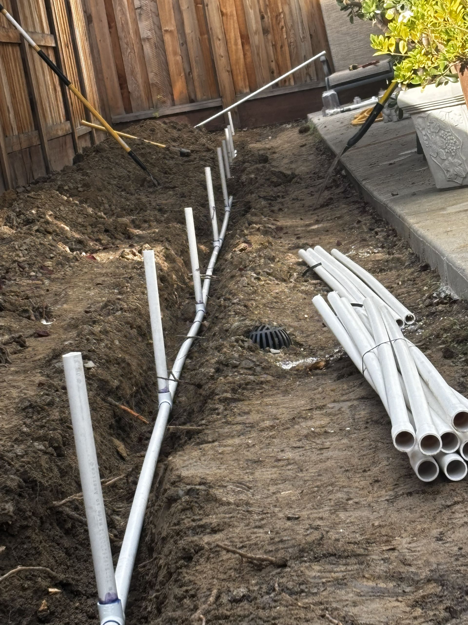 PVC pipes laid along a dirt trench in a backyard, with more pipes stacked on the right.