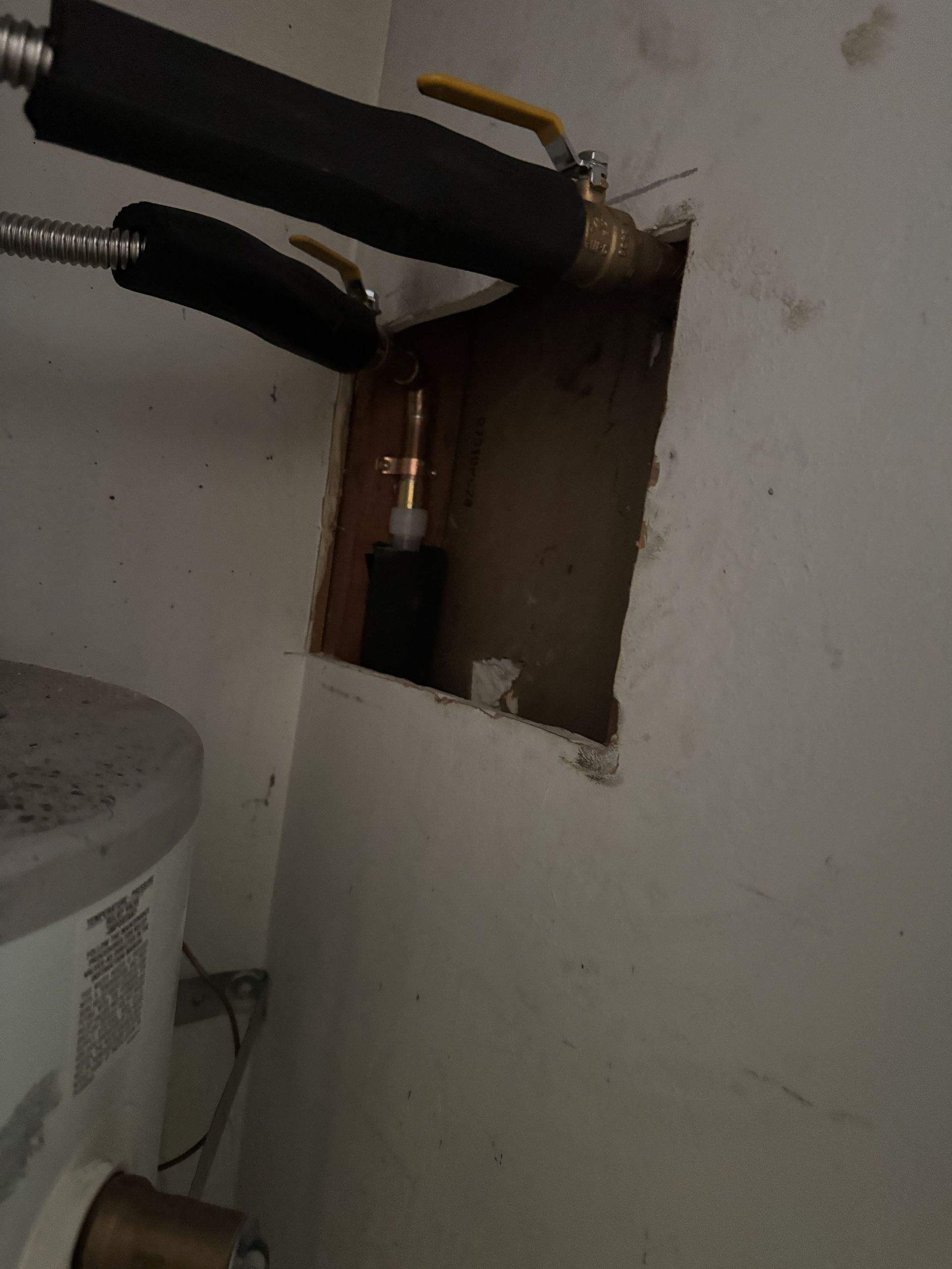 Wall opening exposing pipes and cables beside a white utility unit