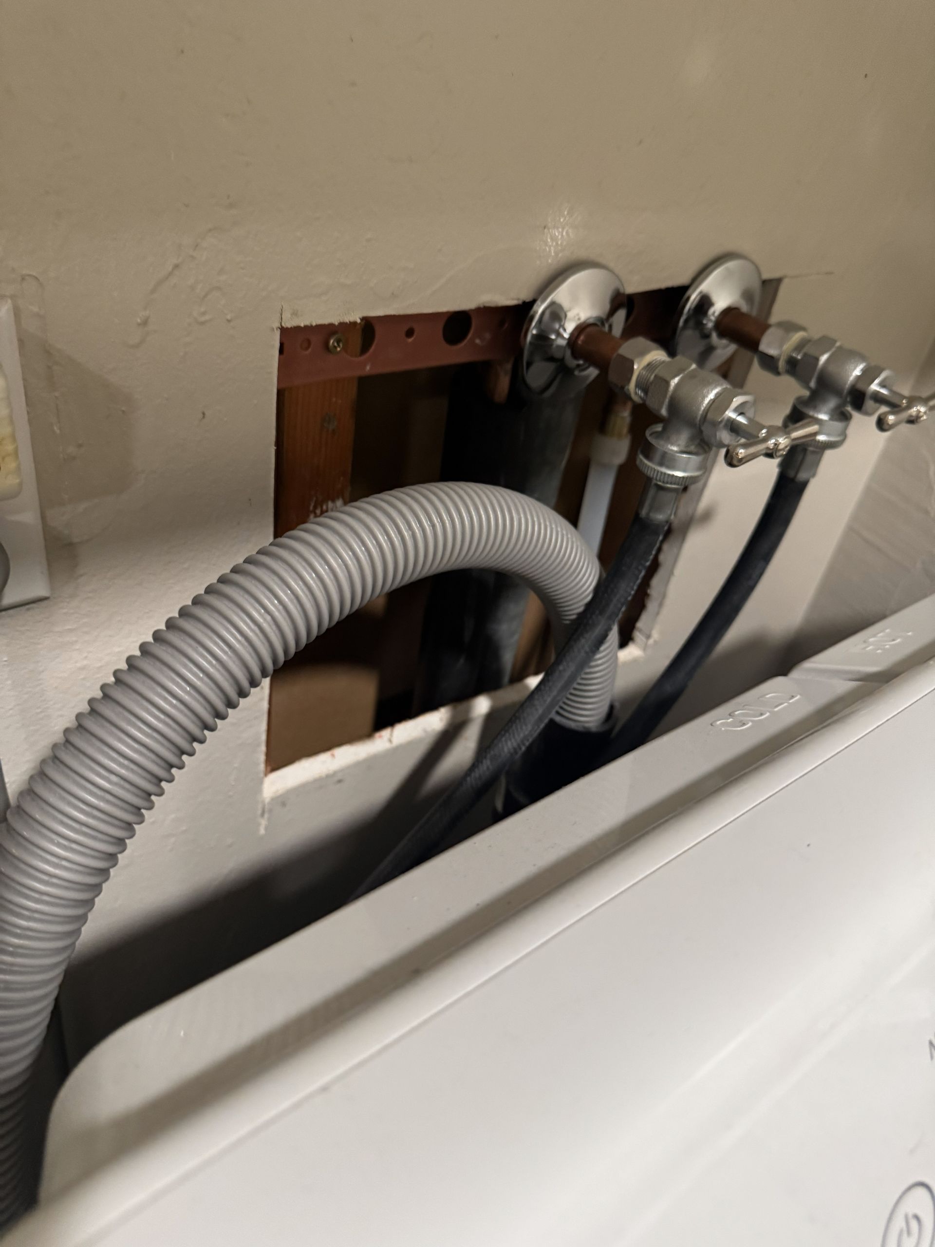 Exposed washing machine plumbing with gray hoses and metal valves above a white appliance