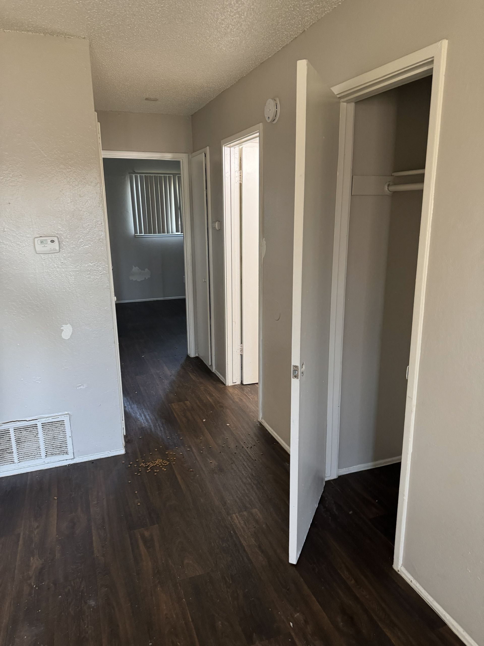 Empty hallway with dark wood floors, white walls, open closet, and a doorway to a brighter room.