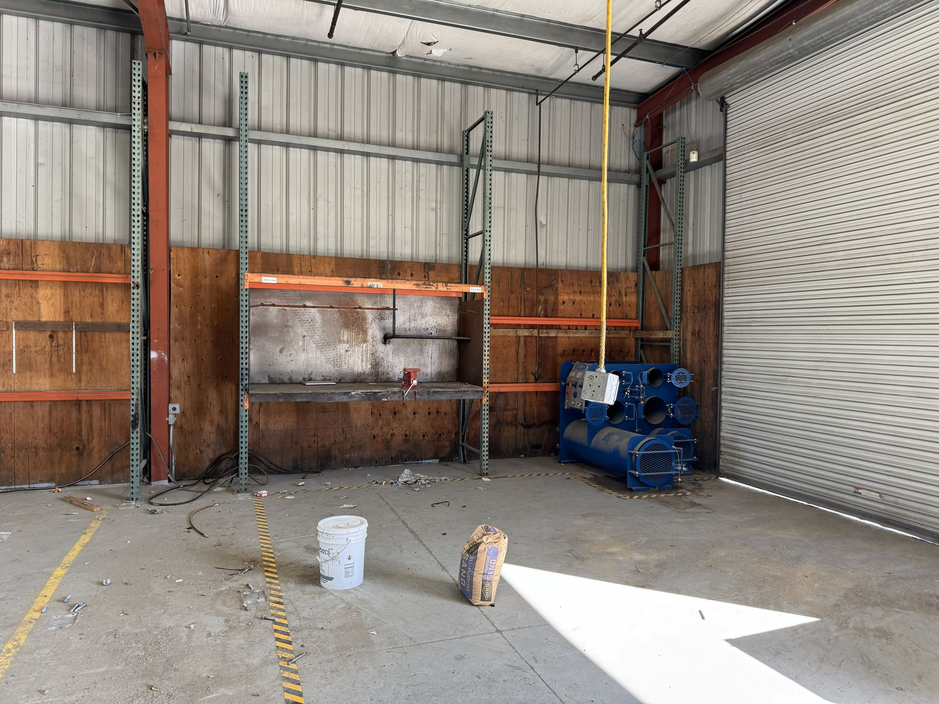 Empty warehouse interior with metal walls, open roll-up door, and a bucket on the concrete floor.