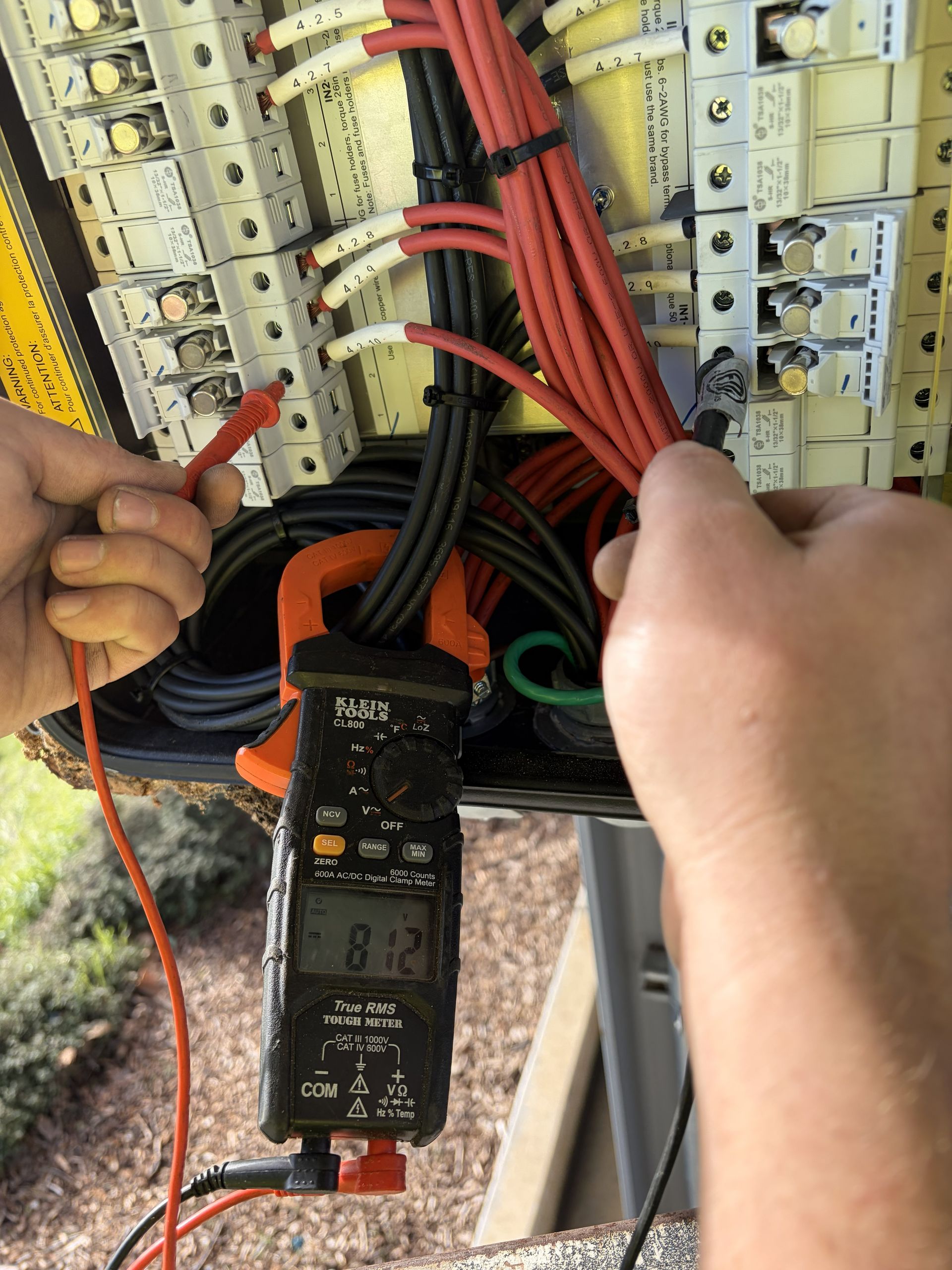 Hands using a clamp meter to test red wires in an electrical panel