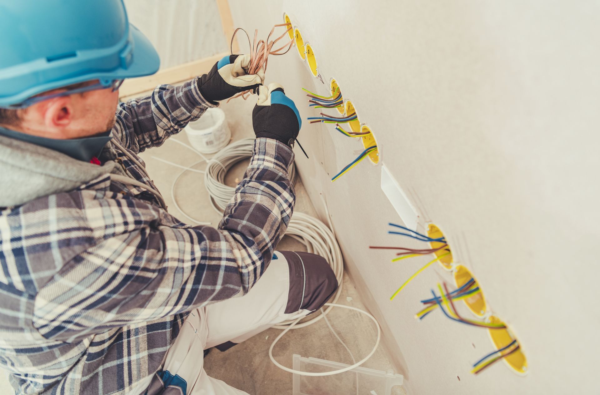 Close-up of a commercial electrician, wearing a blue helmet and dark gloves, installing outlets. Electrician in blue hard hat wiring cables in a wall panel