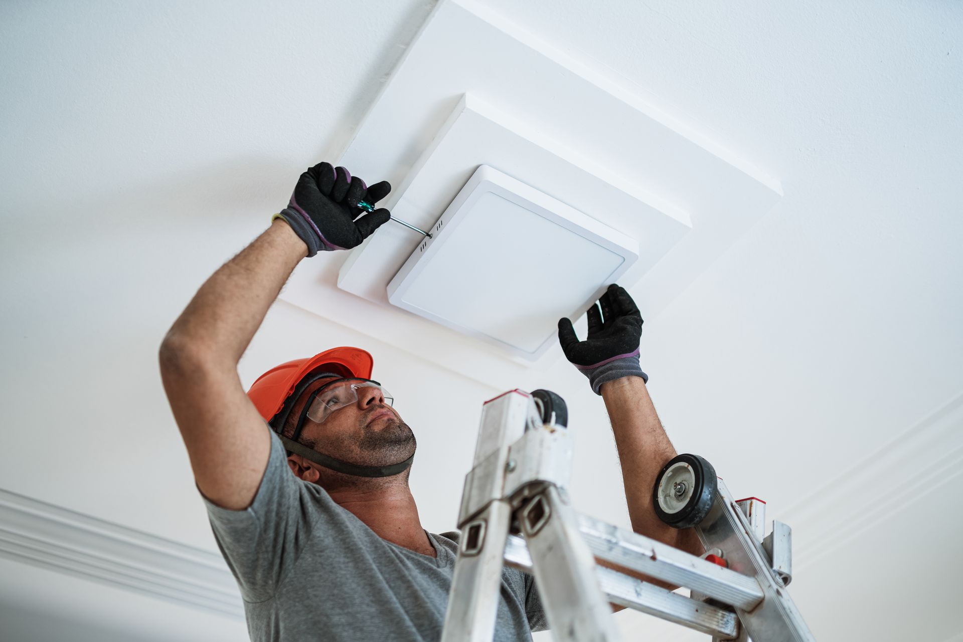 An electrician, wearing a helmet, glasses, and gloves, is installing an LED light on the ceiling. Technician on a ladder installing a ceiling air vent, wearing a red hard hat and black gloves