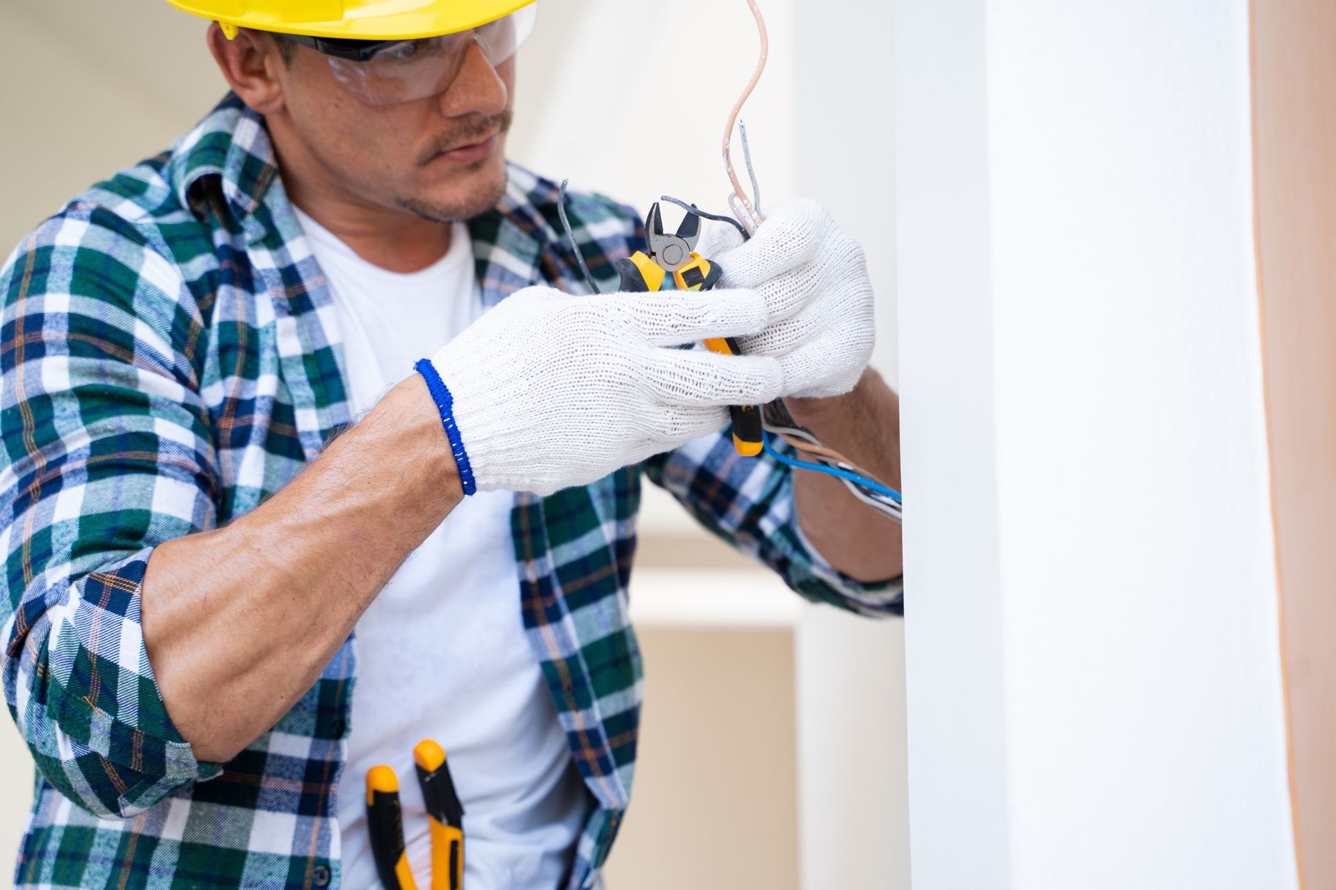 Electrician in a hard hat wiring an outlet inside a wall, wearing gloves and plaid shirt