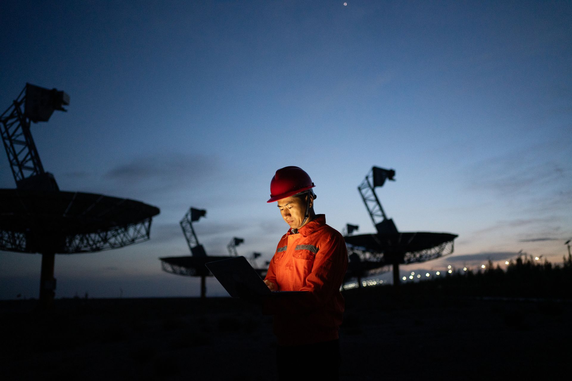 Worker in red hard hat and jacket standing at dusk in a radio telescope array field.