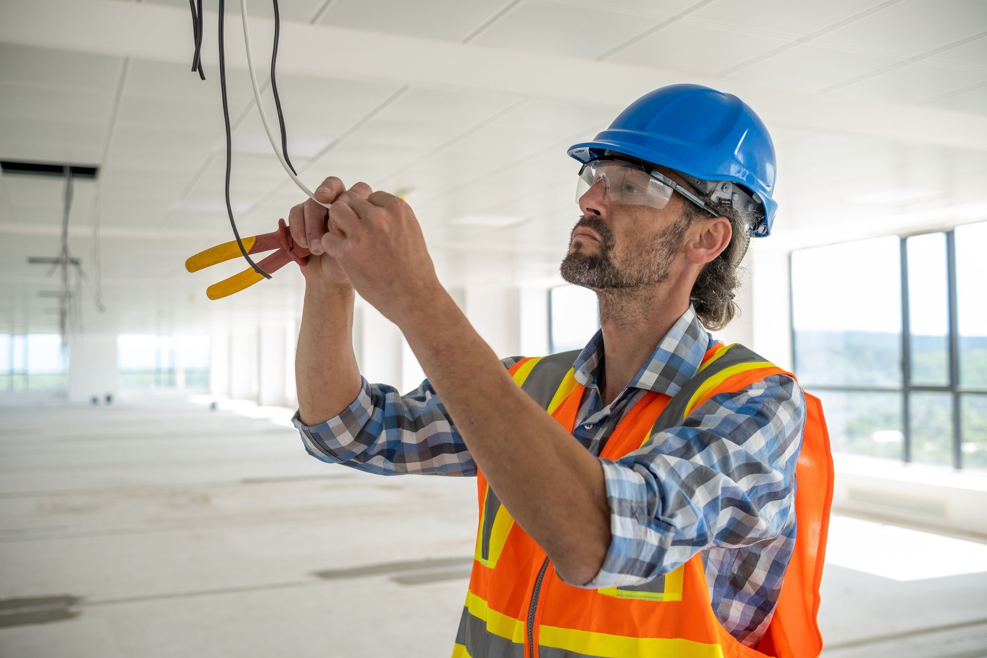 Electrician preparing the cables for the installation of lighting fixtures. Construction worker in blue hard hat testing ceiling wires in a bright unfinished room