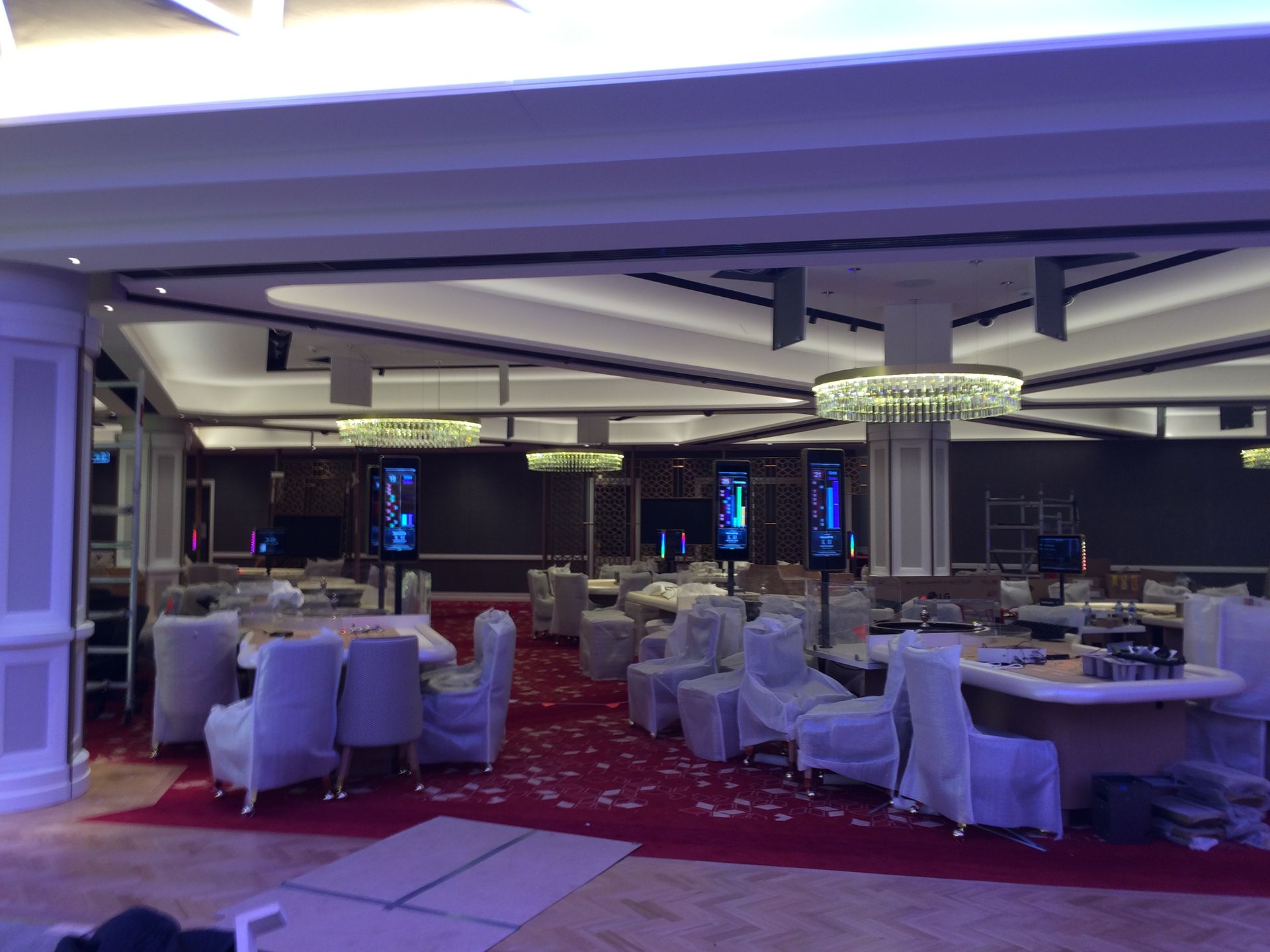 Elegant banquet hall with round, set tables, white chair covers, and red carpet lighting.