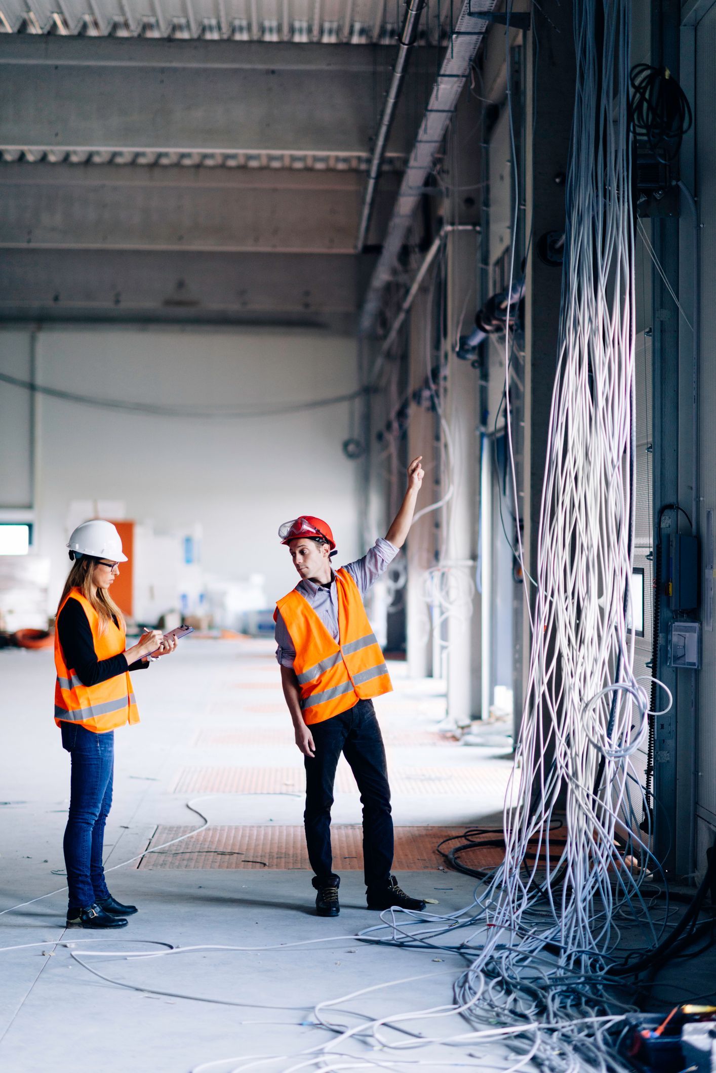 Two construction workers in orange vests and hard hats inspect cables in an industrial interior