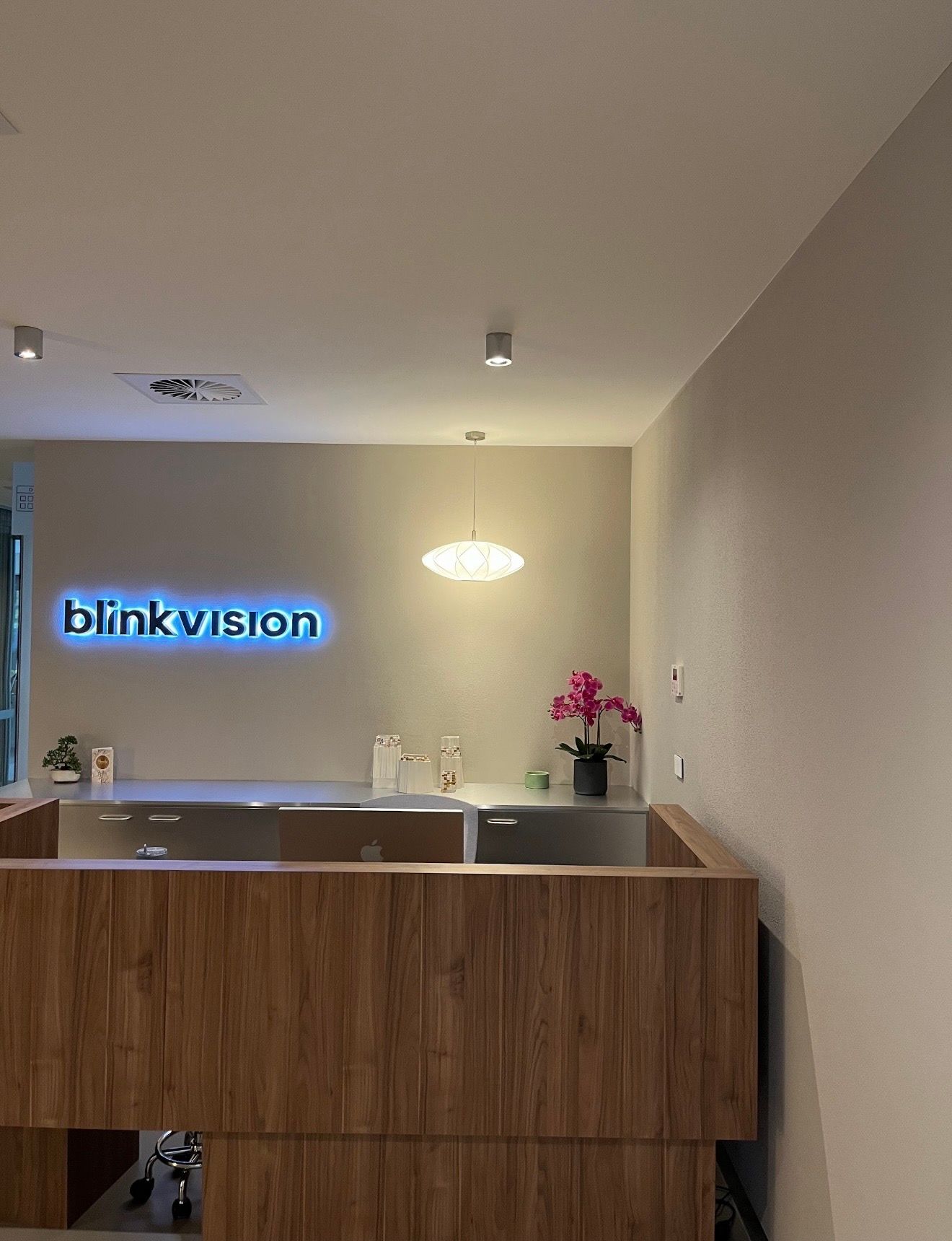 Modern office reception desk with a glowing “blinkvision” sign and pink flowers under a pendant light.
