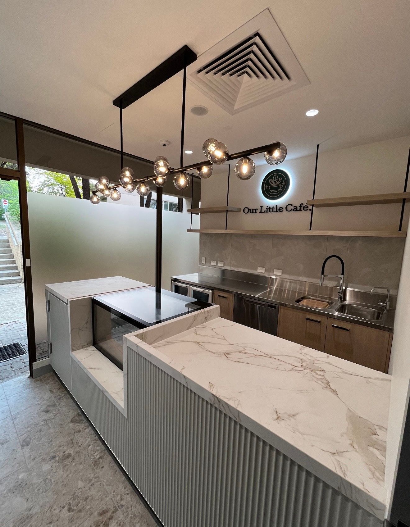 Modern café counter with marble top, pendant lights, sink, and stainless prep area in a bright minimalist interior