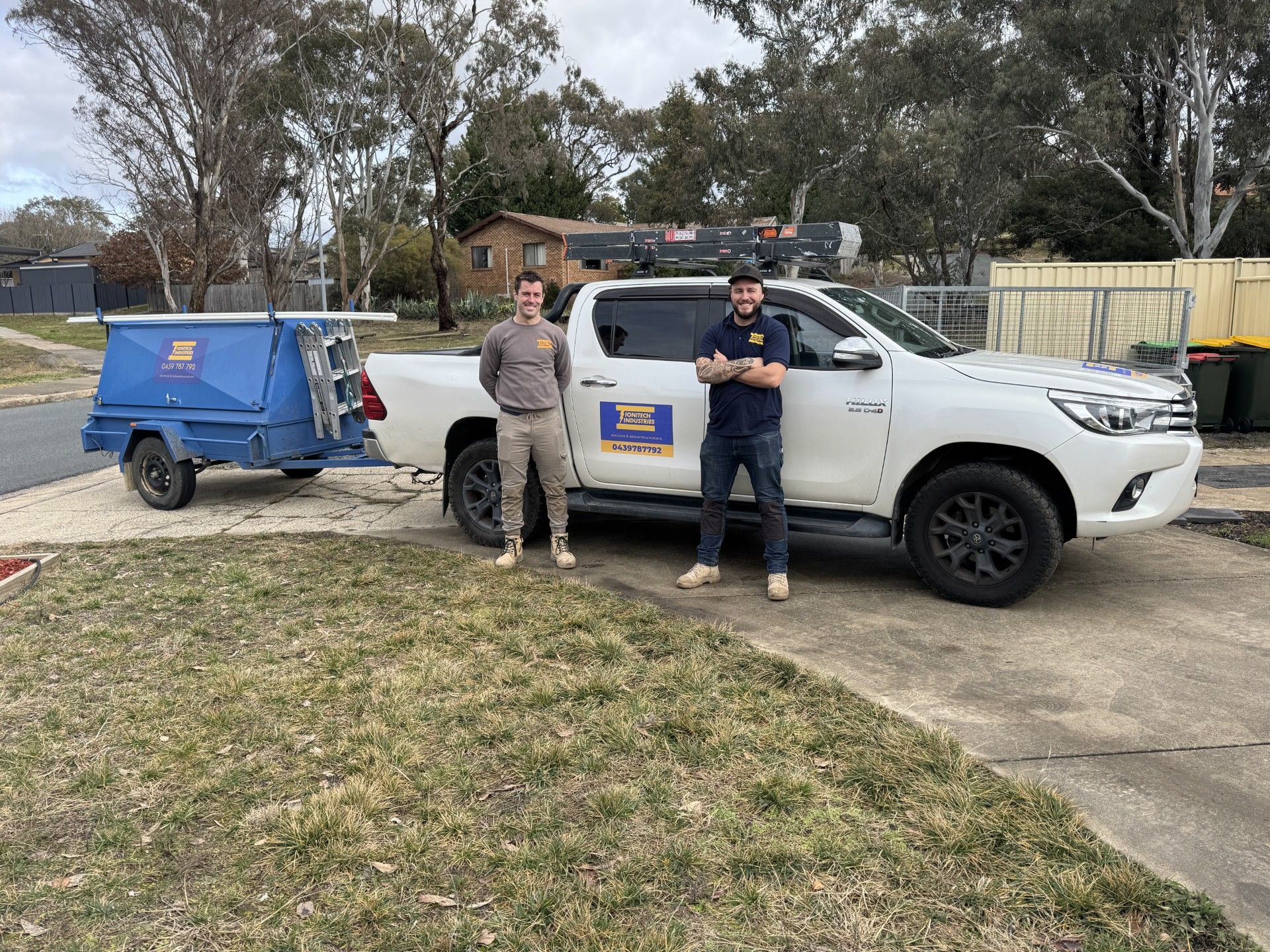 Two people stand beside a white pickup and blue trailer in a driveway, with trees and a fence behind them.