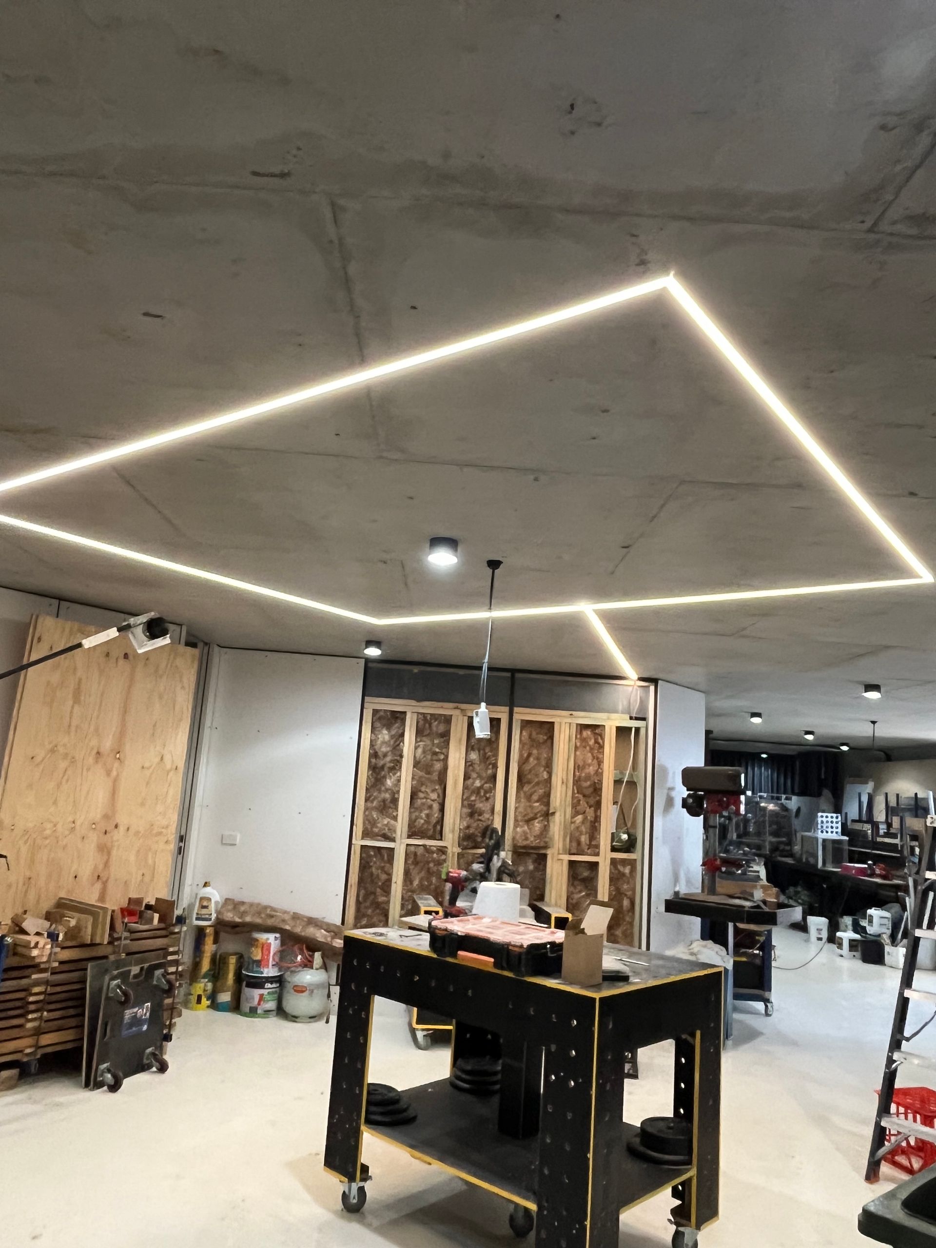 Workshop interior with unfinished ceiling lights, tables, shelves, and exposed wood framing.