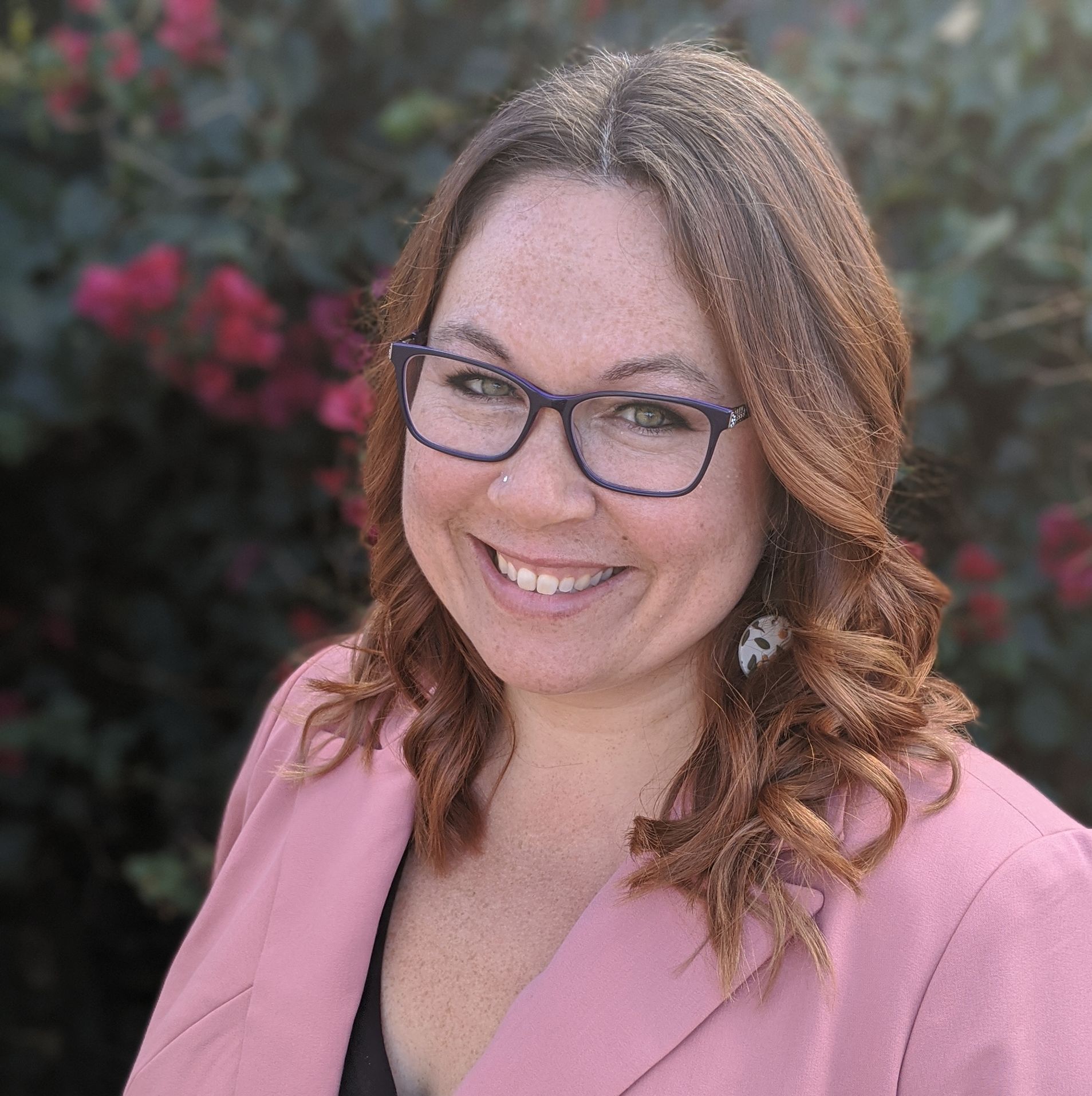 A woman wearing glasses and a pink jacket is smiling for the camera.