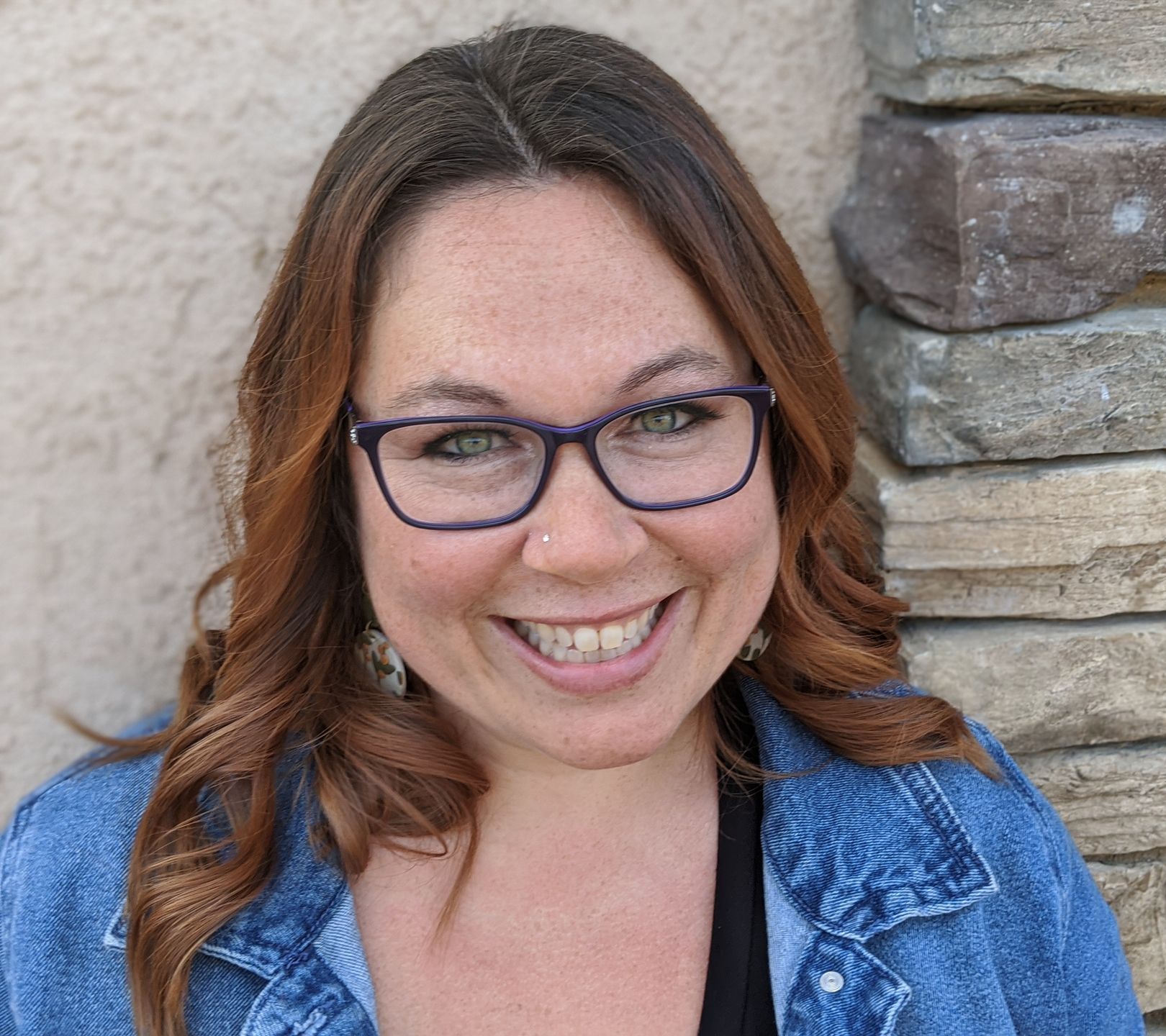 A woman wearing glasses and a denim jacket is smiling for the camera.
