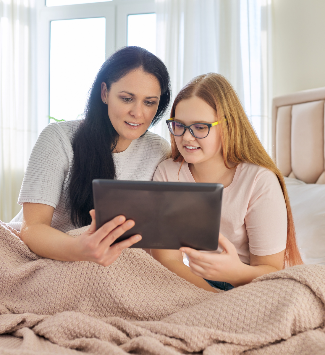 A mother and daughter are sitting on a bed looking at a tablet.