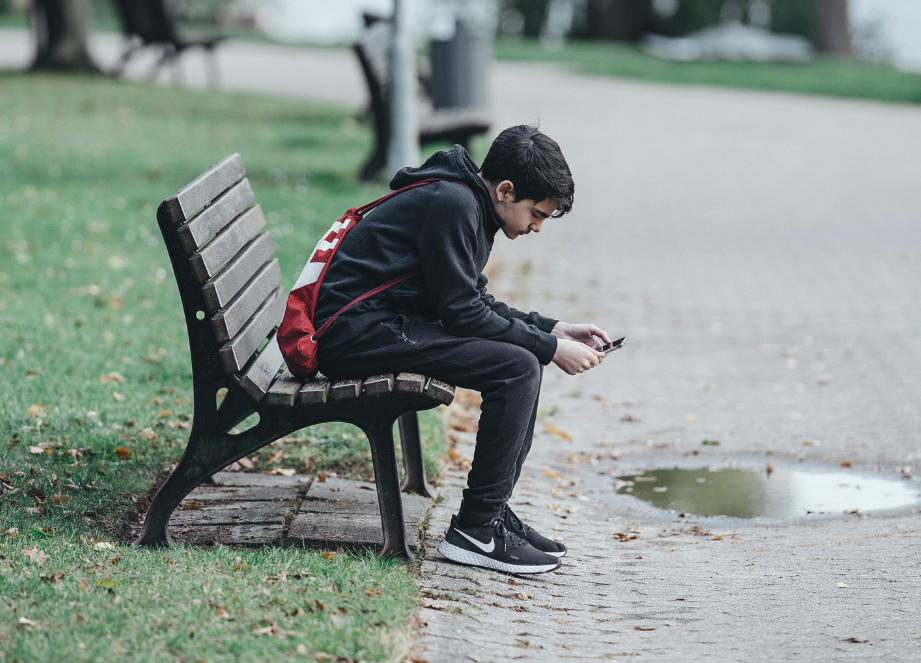 Teenage boy with backpack on sitting on park bench alone looking at his phone.