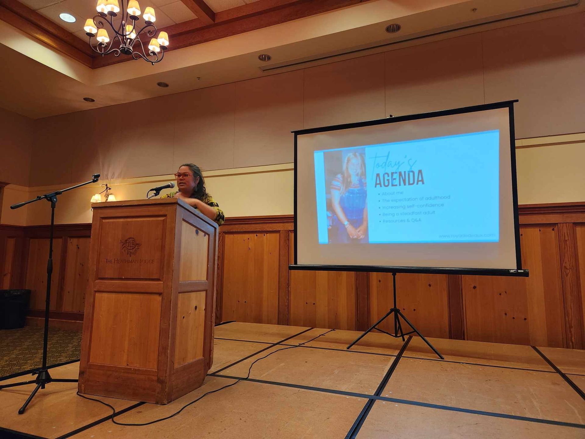 A woman stands at a podium in front of a projector screen that says agenda