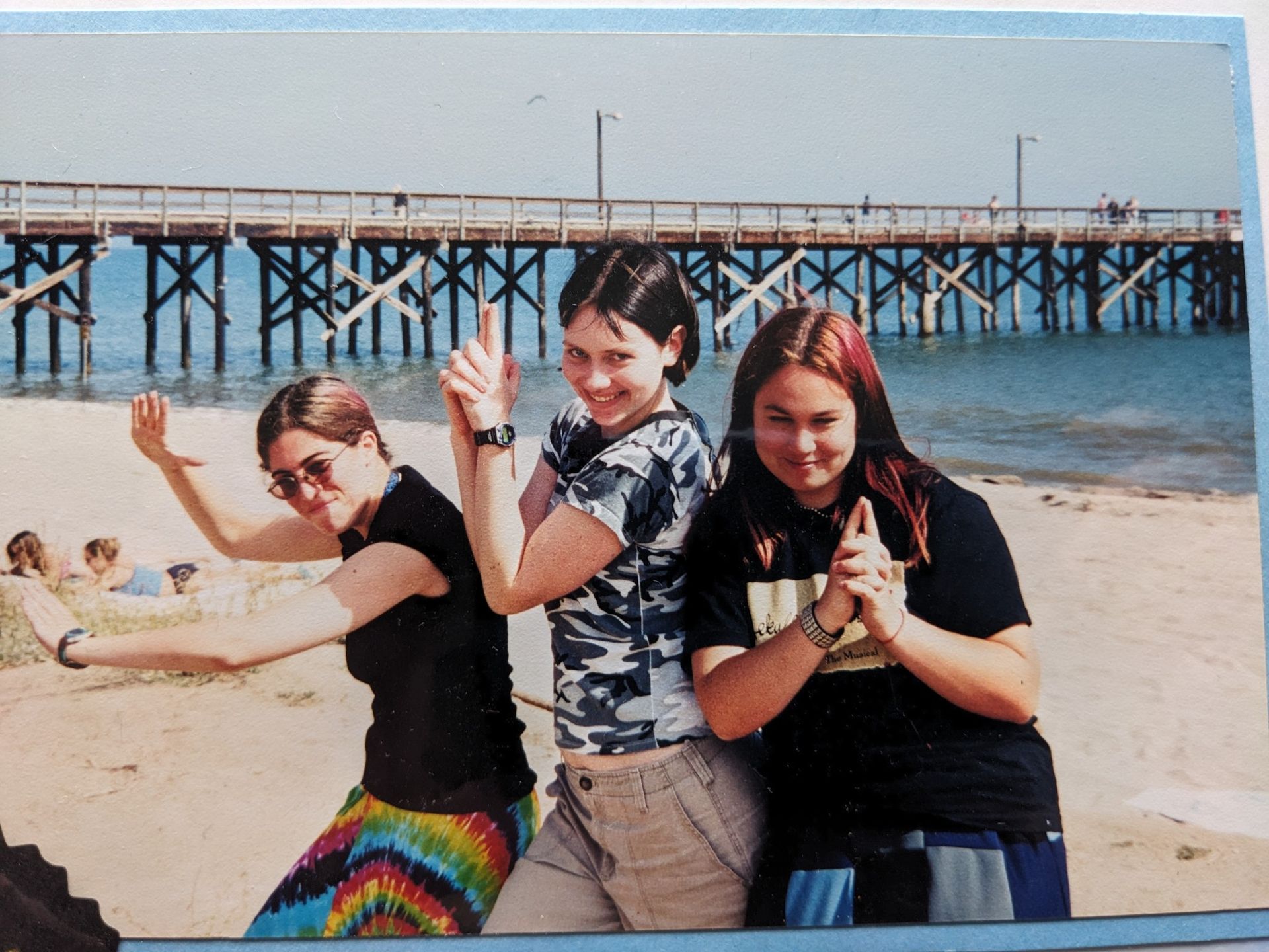 Three girls are posing for a picture on the beach with a pier in the background