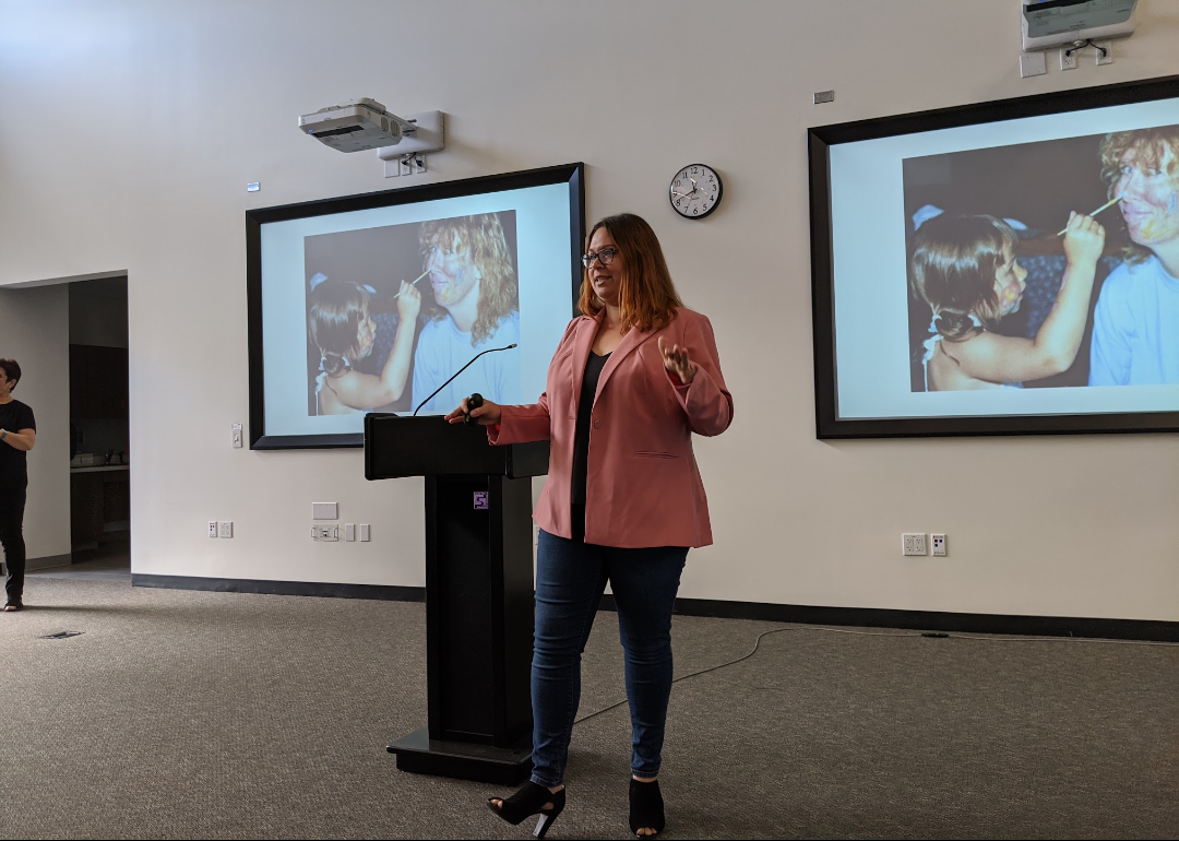 A woman in a pink jacket is standing at a podium giving a presentation.
