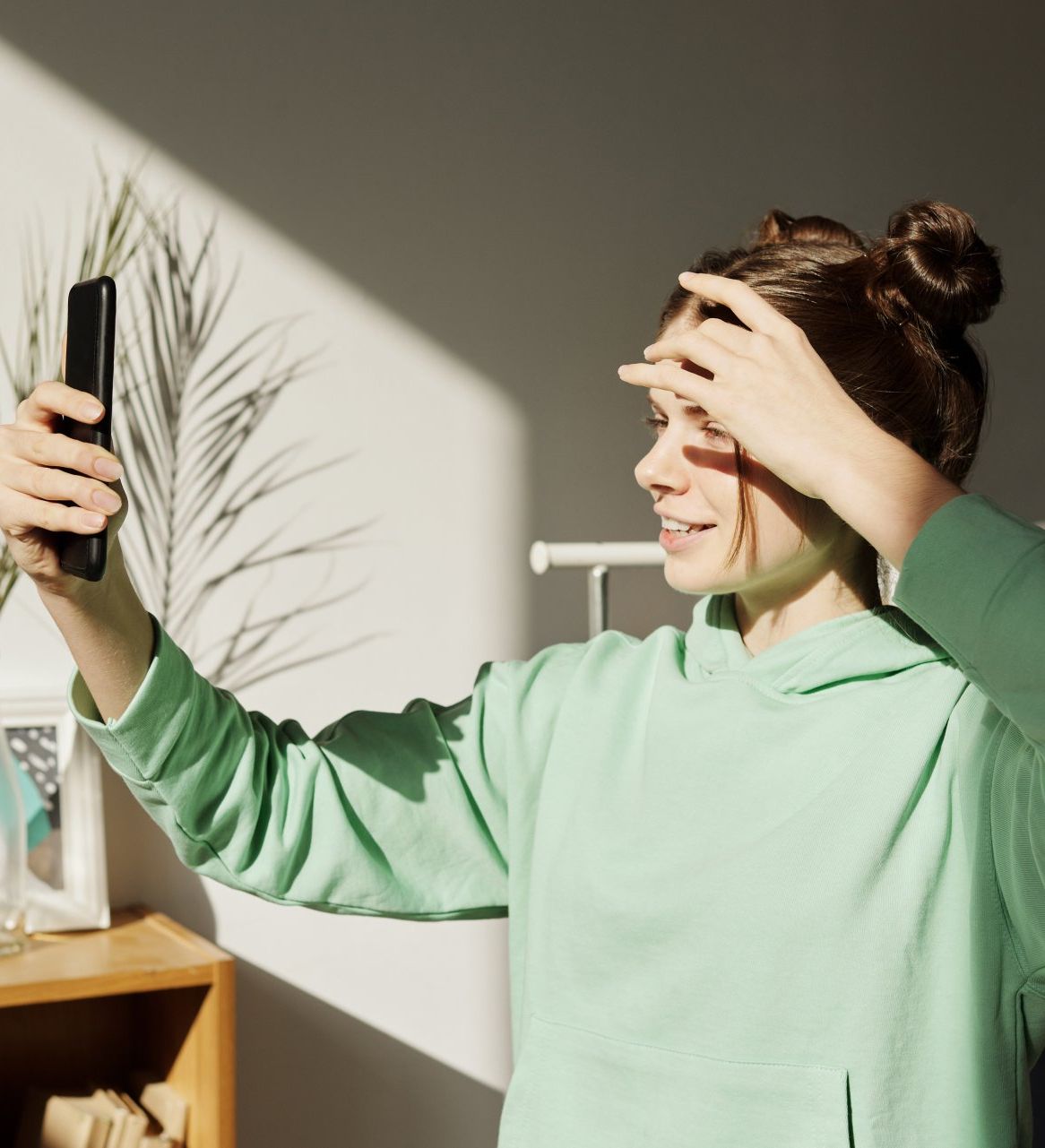 A woman in a green hoodie is taking a selfie with her phone.