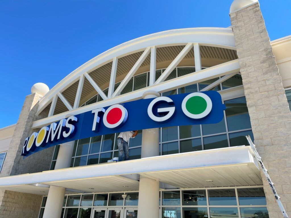 Rooms To Go store exterior with a person working on the sign under a blue sky.